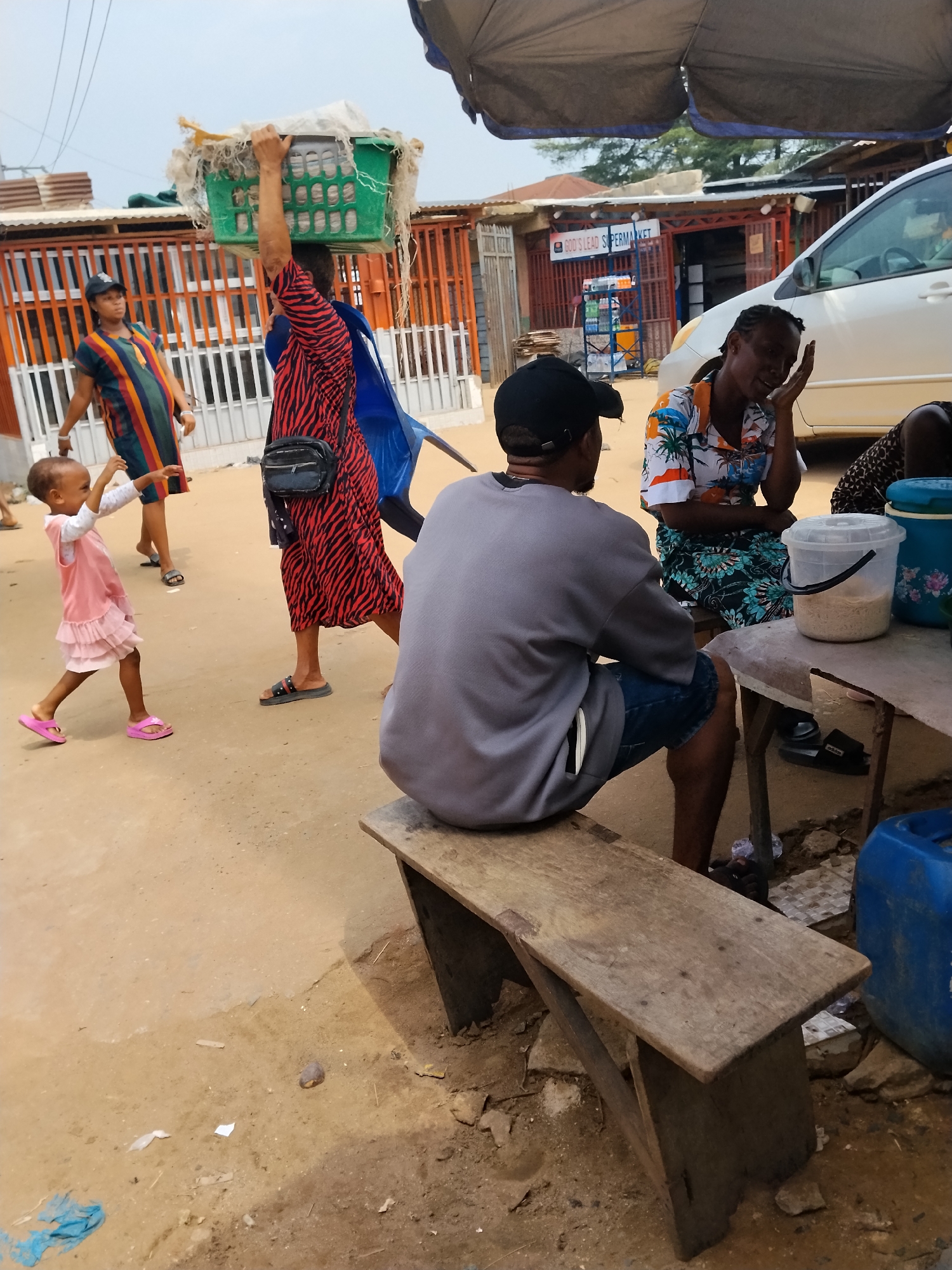 In Ura Nta Umuarandu, Nigeria, a street scene unfolds on a dirt ground. A man in a gray shirt and black cap sits on a worn wooden bench, his back to the viewer. Beside him, a table holds a clear plastic bucket with white granules, alongside blue containers, and two women are seated at the table. One woman wearing a patterned shirt touches her face, and a second woman sits beside her.
To the left, a small child in a pink dress and sandals raises her hands, seemingly interacting with a woman in a colorful striped dress walking nearby. Another woman in a red and black zebra-striped dress carries a green basket on her head, with a black purse slung across her body. A blue material trails down her back.
Behind them, a building with an orange gate is visible. In the background, a white car is parked near a store front labeled "GOD'S LEAD SUPERMARKET." A large umbrella provides shade over the table where the man and women are sitting.