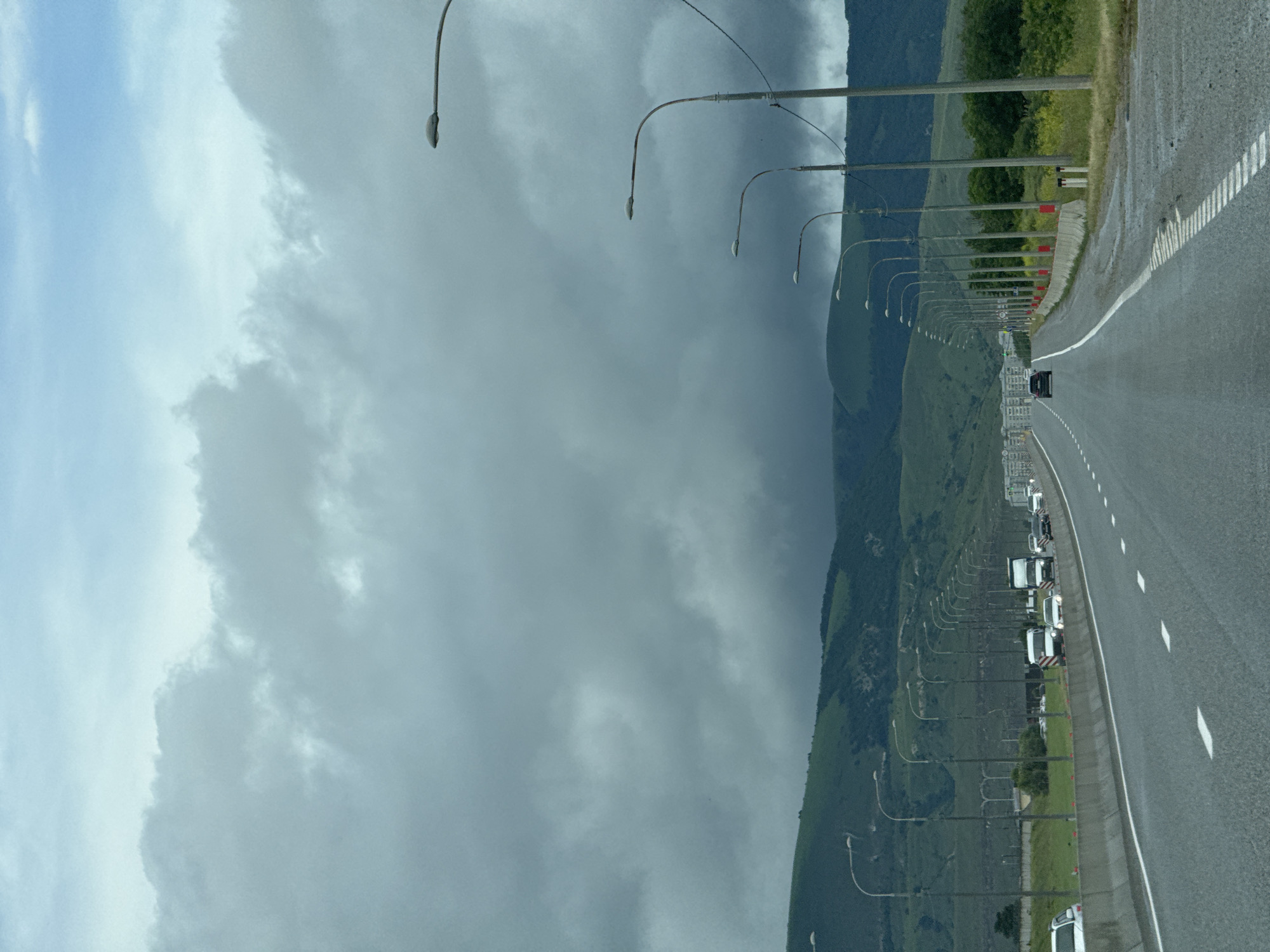 The image shows a highway with multiple lanes, likely in a suburban or rural area. The sky is overcast with dark, heavy clouds, suggesting an impending storm or rain. A line of streetlights runs along the right side of the highway. In the distance, there are green, rolling hills covered in vegetation. Several vehicles, including cars and trucks, are visible on the highway. The road surface appears wet, indicating recent rainfall or damp conditions. There is no visible signage or context that specifically identifies the location as Красногорская, Russia.