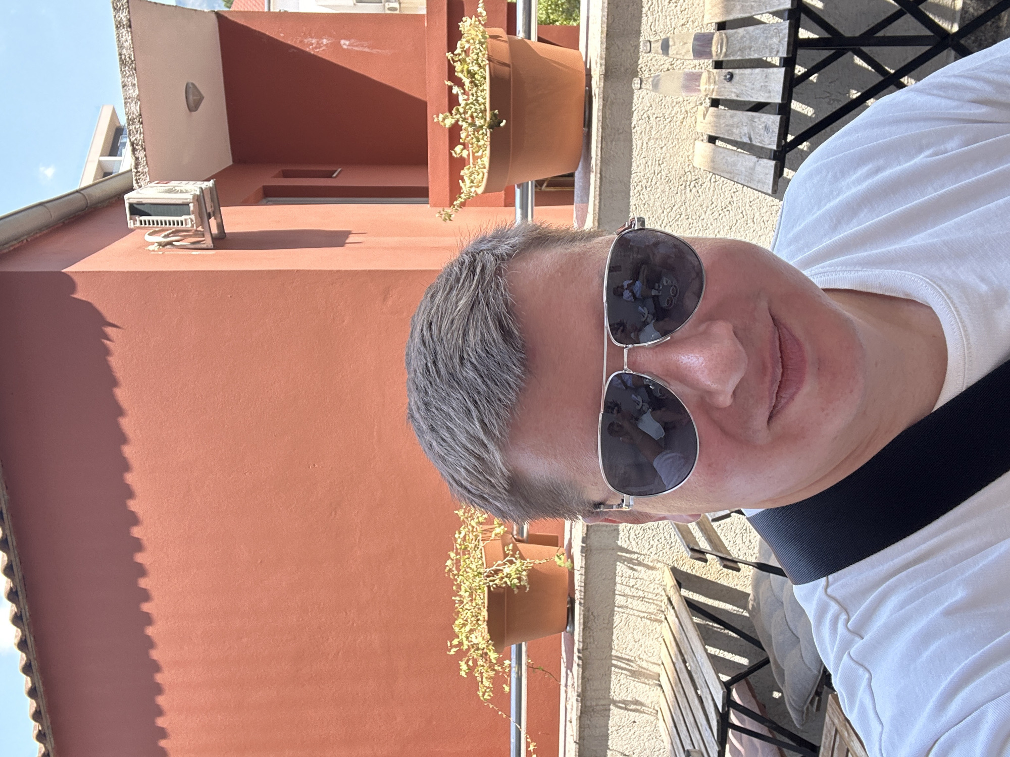 The media file is a close-up, rotated selfie of a person wearing sunglasses and a white t-shirt. The subject has short, graying hair and appears to be outdoors. In the background, there is a terracotta-colored building with a balcony adorned with hanging plants. A metal railing and a textured wall are also visible. The sunlight suggests it is daytime. The sunglasses reflect a scene that includes other people and possibly a table. The overall impression is of a casual moment, likely a vacation or leisure activity, given the outdoor setting and relaxed attire. There is no visible text in the media file. The location context of Budva, Montenegro, suggests a coastal European setting, possibly with a Mediterranean architectural style.