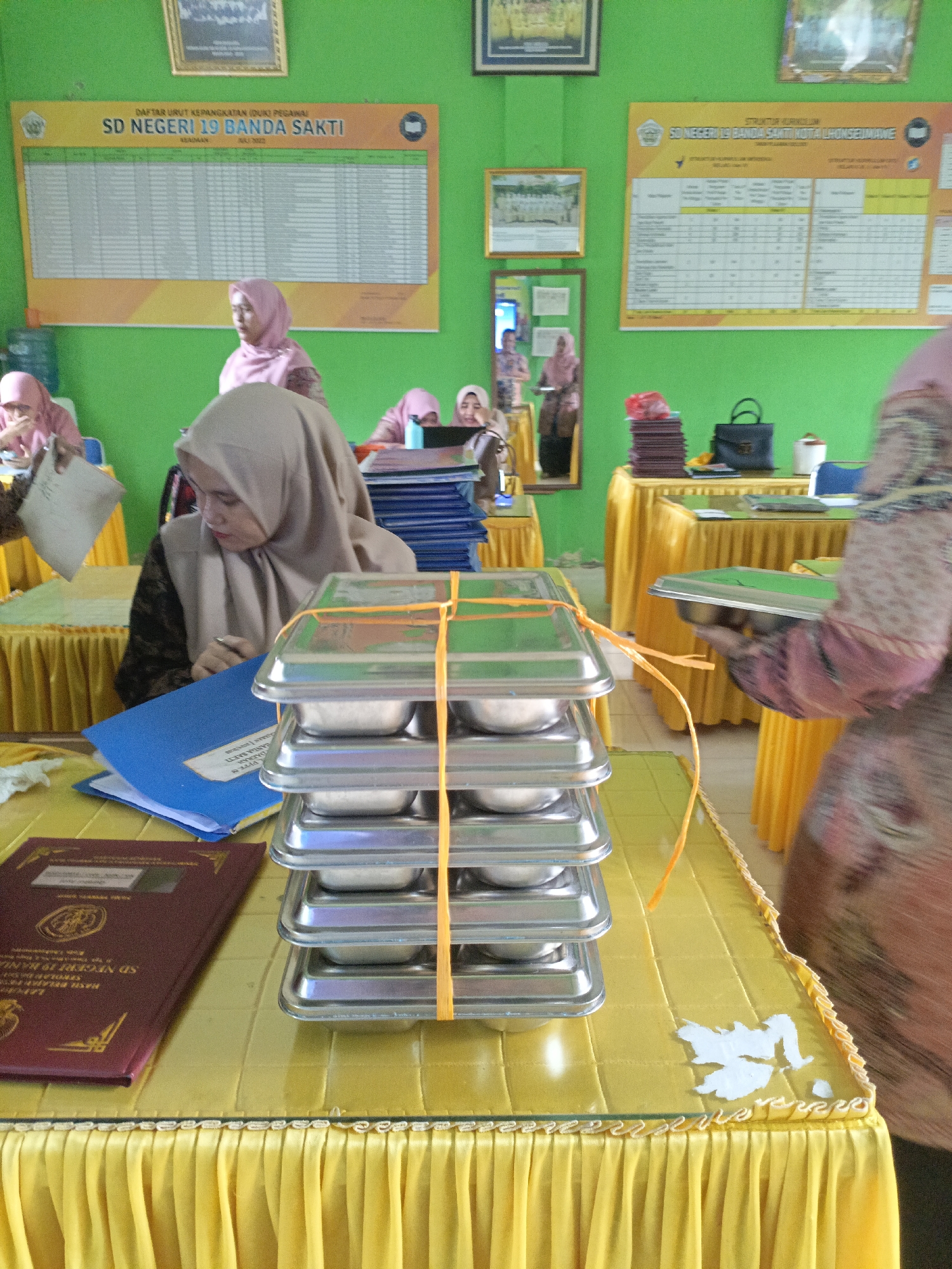 In Lhokseumawe, Indonesia, a classroom scene is depicted with predominantly green walls. Several individuals wearing hijabs are present. In the foreground, a stack of silver food trays, bound by an orange string, is placed on a yellow-draped table. To the left, a woman wearing a beige hijab is looking down and writing on a blue folder. Another person's hand is visible holding a document. On the table, a maroon folder with gold emblem and text is also present. In the background, a bulletin board displaying rows of text is mounted on the wall. Another bulletin board is visible further back, also featuring charts and text. To the right, a woman in a patterned hijab and dress is holding a silver food tray, seemingly in the process of serving. Tables draped in yellow fabric and chairs are distributed throughout the room.