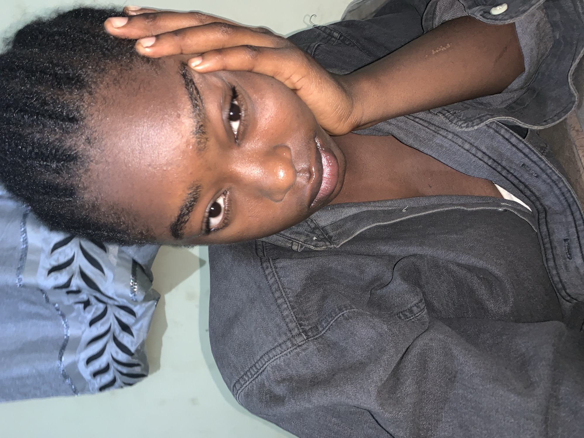 A close-up, high-angle shot shows a young Black person reclining. The person is wearing a dark, buttoned denim shirt or jacket. Their head is resting on their hand, with their fingers touching their forehead. They are looking directly at the camera. The background includes a blue-gray patterned pillow. There is no evidence to confirm location in Rafin Par, Nigeria.