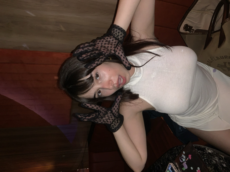 A young woman poses in a white dress and black lace gloves. She holds her hands near her face, giving a playful expression. The background is dimly lit, suggesting an indoor setting. A designer tote bag rests near her, and part of a table is visible in the foreground. The image is taken from a low angle.