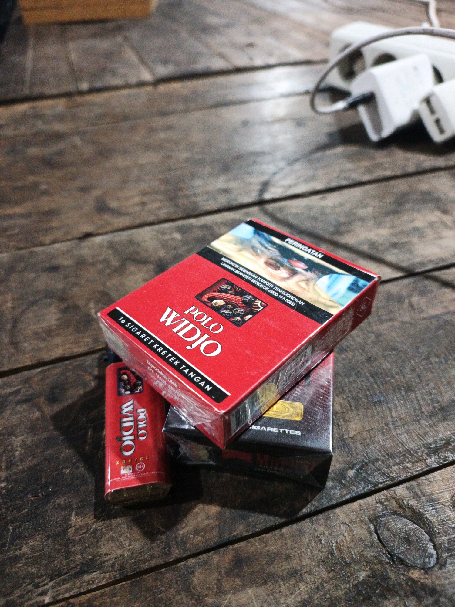 A close-up shot, taken from a slightly elevated angle, shows three packs of cigarettes arranged on a weathered wooden surface. The dominant pack, positioned at the top and angled to the right, is bright red and displays the brand name "POLO WIDJO" in large white lettering. Smaller text below reads "16 SIGARET KRETEK TANGAN". The upper portion of this pack features a graphic warning with a disturbing image and text in Indonesian.
Beneath the red pack, two other cigarette packs are visible, partially obscured. The pack directly below appears to be black and silver with the word "CIGARETTES" visible, and some text in what seems to be a foreign language. A smaller, red cigarette pack, with "POLO WIDJO" branding, lies to the left of the main red pack, leaning against it. This smaller pack has a warning label indicating "18+" and "PERINGATAN".
The wooden surface exhibits a rough texture with visible grain, cracks, and knots, suggesting an outdoor or rustic setting. In the background, out of focus, a white electrical extension cord with multiple outlets and a plug is visible on the wooden surface, indicating the presence of modern electrical accessories. The lighting suggests an indoor setting with diffused natural or artificial light. The overall scene is static, with no people or apparent activity. The image does not provide any specific indication of its location within Ketegan, Indonesia.