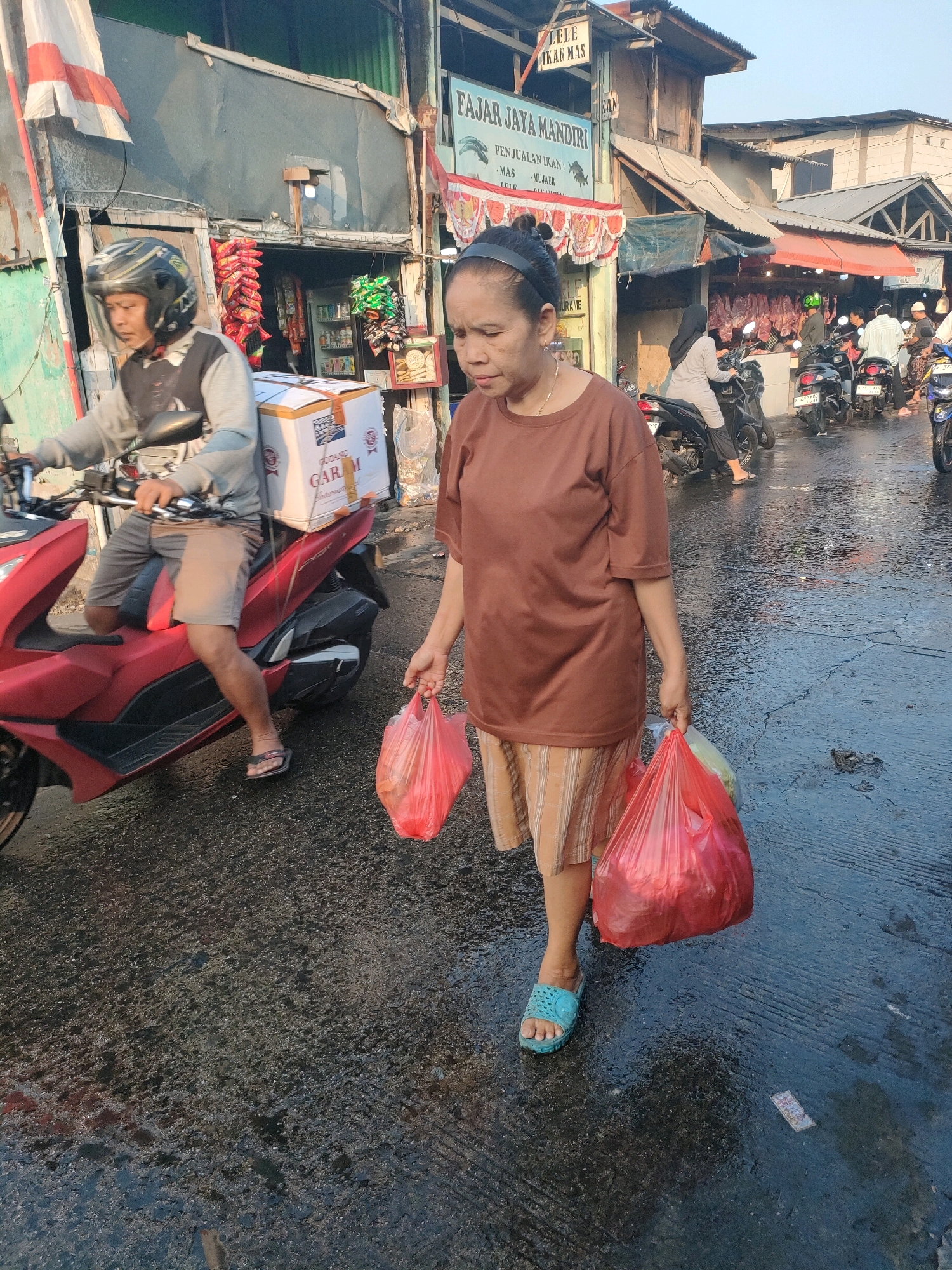 In the city of Bekasi, Indonesia, a middle-aged woman walks across a wet street, holding two red plastic bags filled with groceries. She is wearing a brown t-shirt and a patterned skirt, with blue sandals on her feet. A man on a red motorcycle, wearing a helmet and shorts, is seen in the background. The street is lined with market stalls, displaying various goods including meat and snacks. The scene appears to be a busy marketplace, with other people and motorcycles also present. The atmosphere is vibrant and bustling, suggesting a typical day at the market.