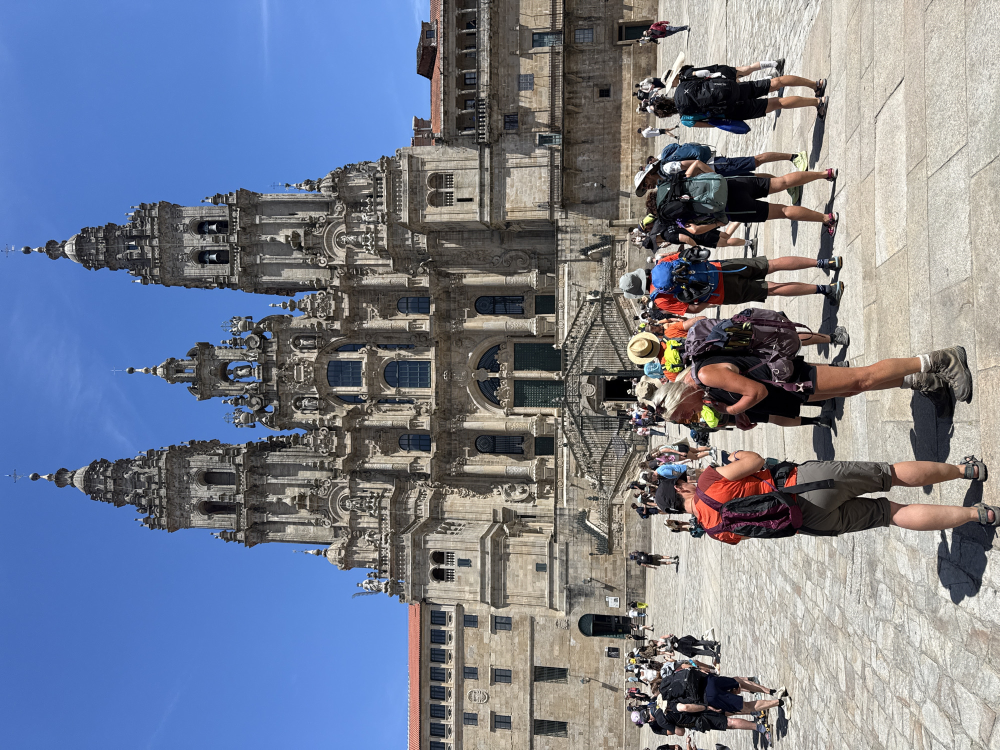 The image shows a sunny day in the Praza da Quintana in Santiago de Compostela, Spain, in front of the Cathedral. A crowd of people, many appearing to be pilgrims with backpacks, are gathered in the plaza. The architecture of the Cathedral, with its ornate stone facade and towers, dominates the background. The ground is paved with cobblestones. People are standing, walking, and interacting with each other. The scene is bustling with activity under a clear blue sky.