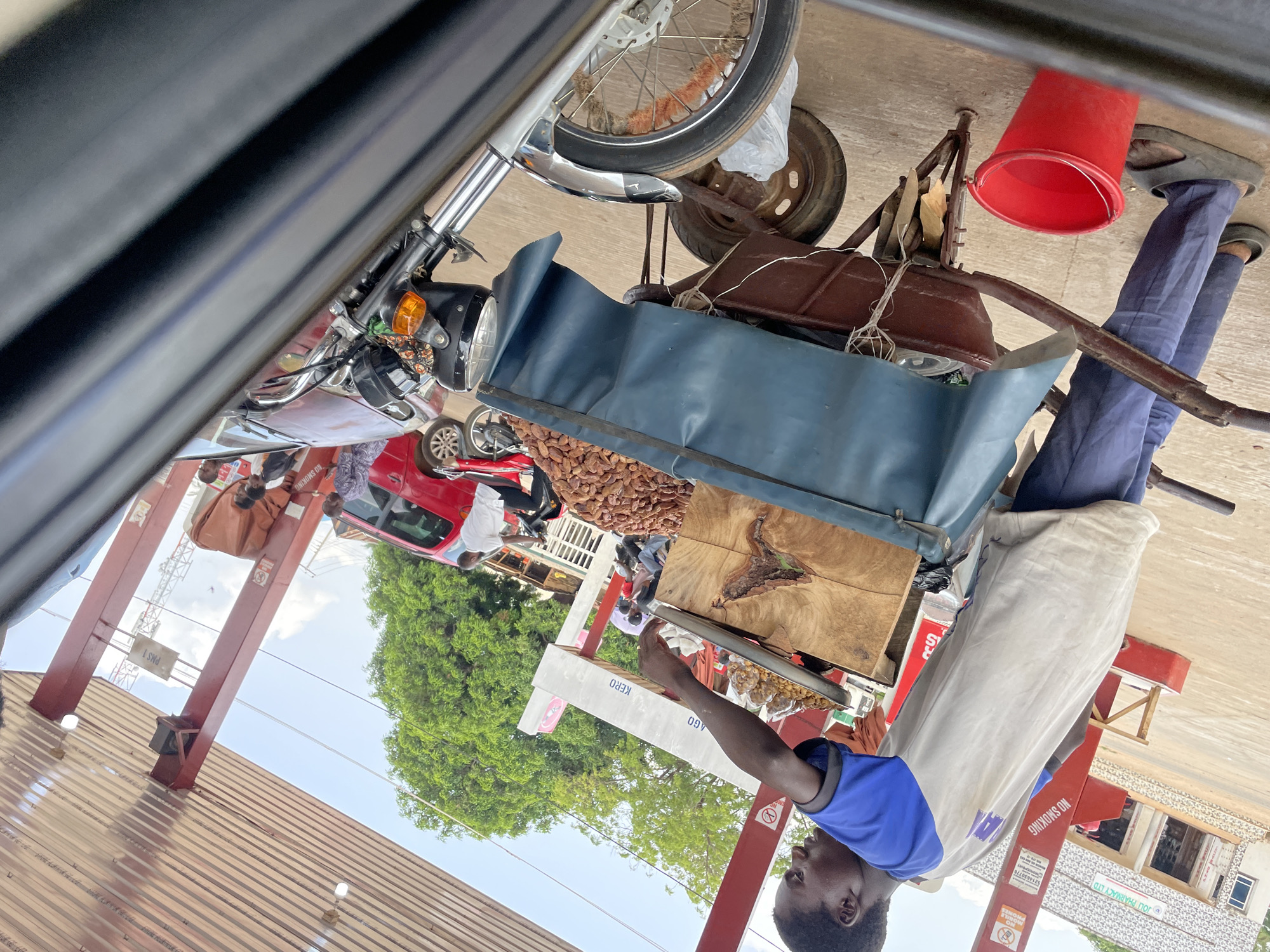 The image shows a street scene in Akwanga, Nigeria. A man in a blue shirt and light-colored pants stands beside a cart laden with goods for sale. The cart appears to be made of metal and wood and has a blue covering over some items. A red bucket is visible on the cart. On the wooden surface of the cart, there are piles of what appear to be dates. The man is holding a metal pan of what might be groundnuts. The cart is parked on what looks like a covered walkway or porch of a building. In the background, there is a red car and other people. A sign reading "NO SMOKING" is visible near the cart. A motorbike is also partially visible.