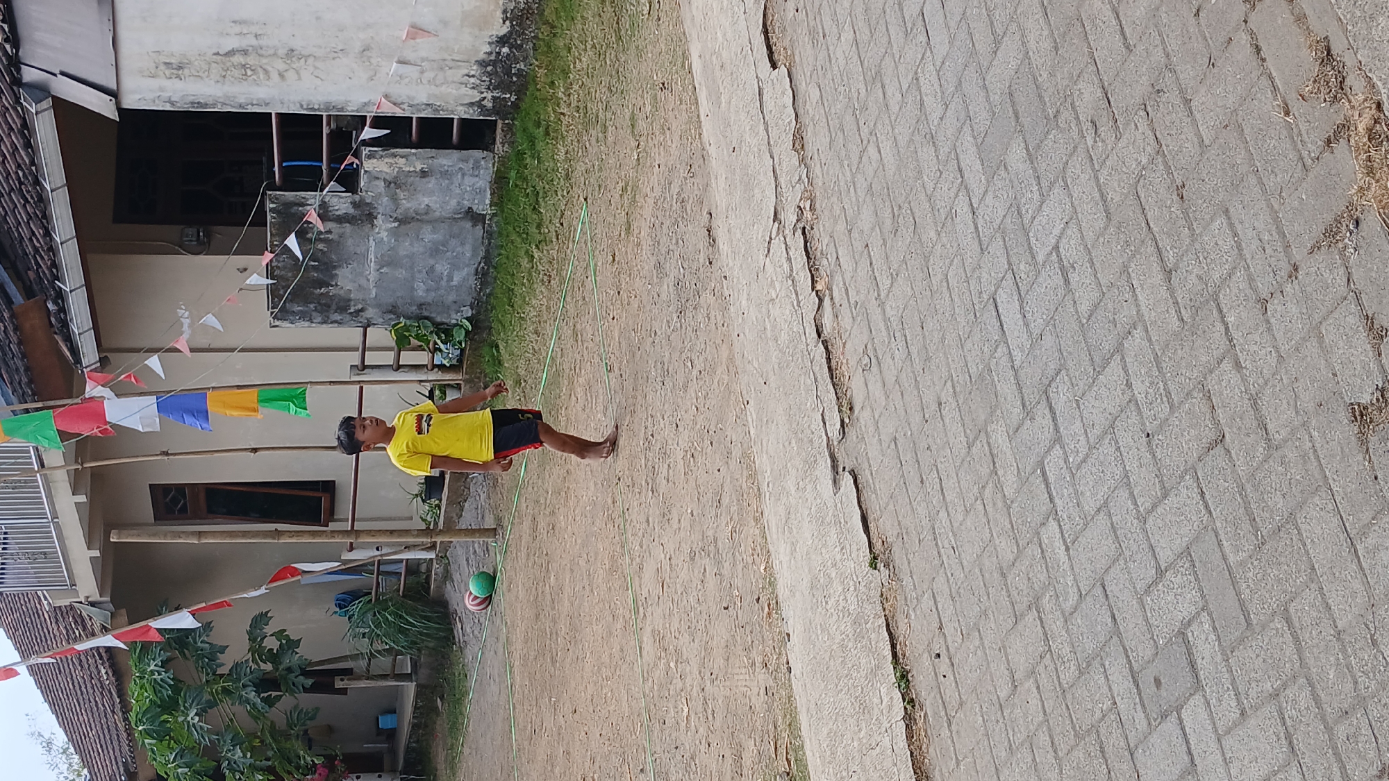 A young boy with dark hair, wearing a yellow t-shirt and black shorts with red accents, walks barefoot across a dirt court. He is in the city of Mendut, Indonesia, with a house visible in the background. The house has a tiled roof and is adorned with Indonesian flags and colorful triangular pennants strung on bamboo poles. Two balls, one red and one green, lie on the court near a thin green line that marks a boundary. The ground in the foreground is paved with interlocking gray bricks.
