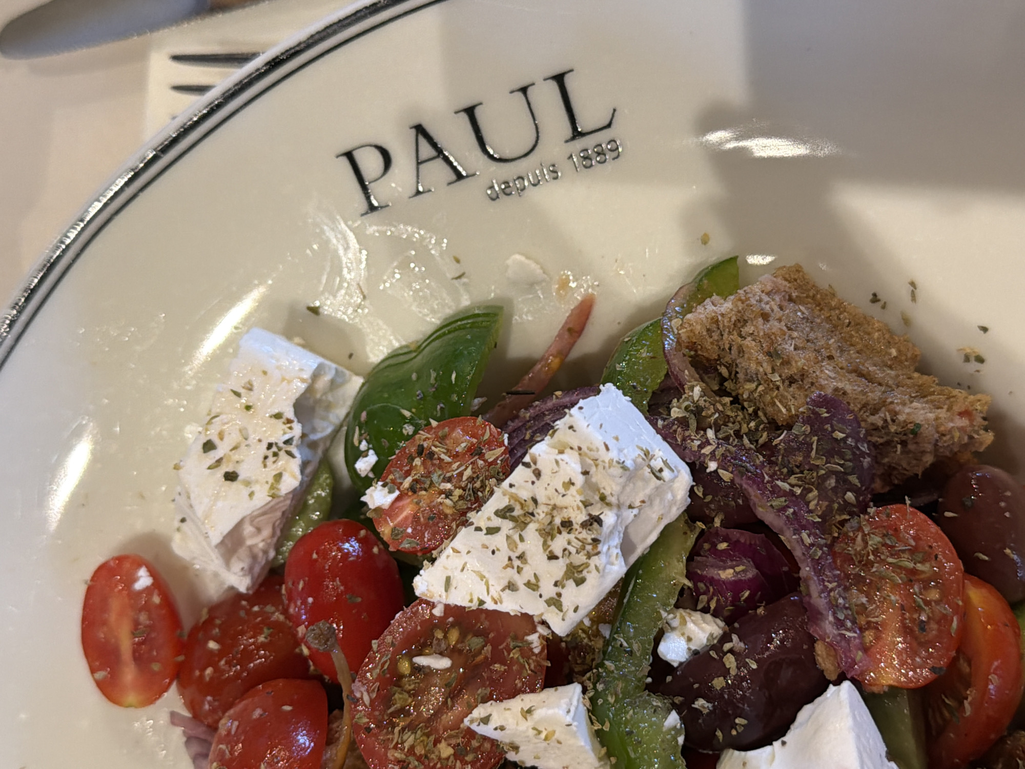 A close-up, overhead shot of a Greek salad on a white plate with a black rim. The salad contains cherry tomatoes, feta cheese, olives, red onion, green bell peppers, and a piece of toasted bread. Dried herbs are sprinkled over the salad. The plate has a logo that reads "PAUL depuis 1889." The setting is indoors, likely a restaurant, with a blurred background.