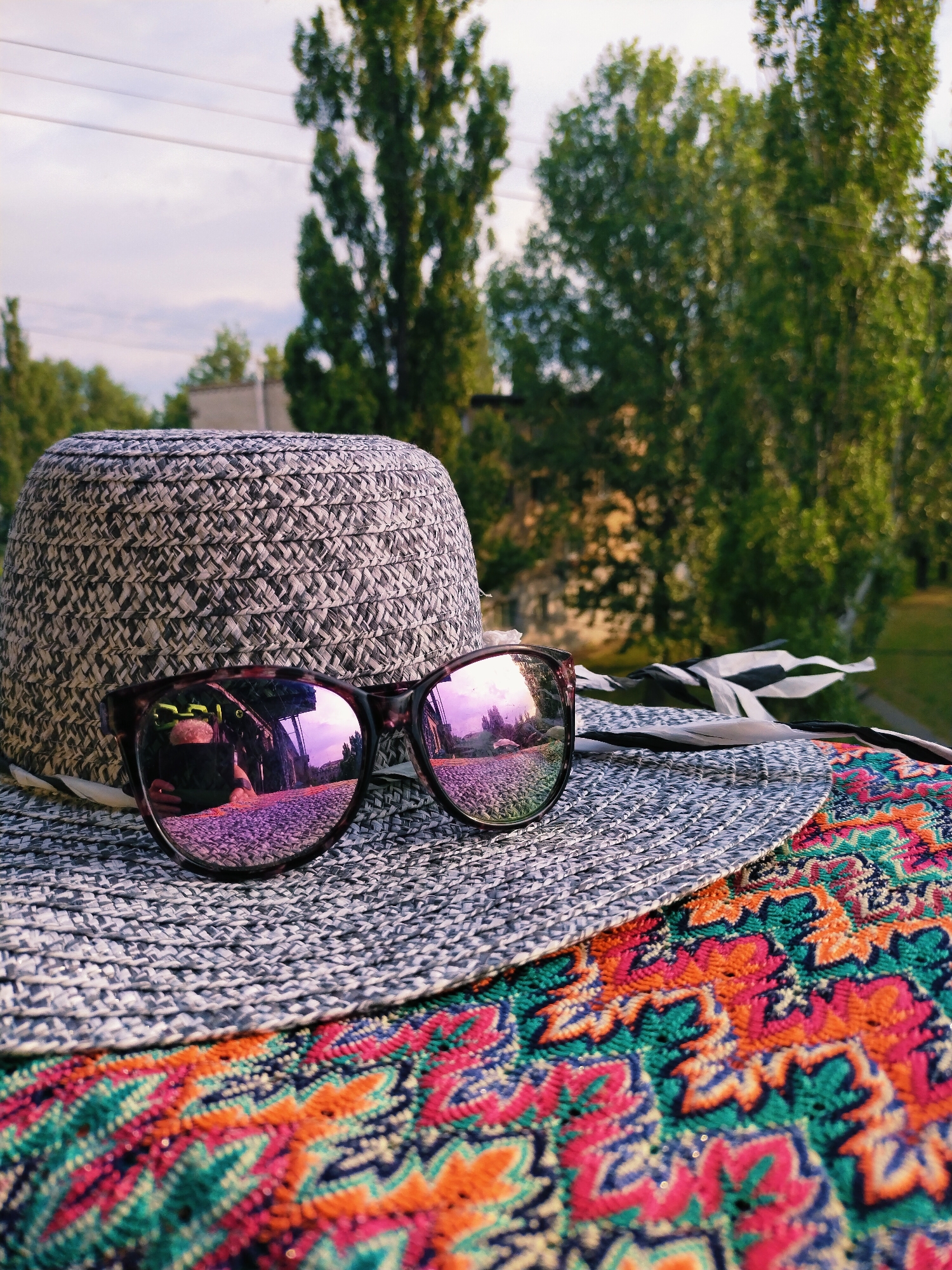In Horishni Plavni, Ukraine, a close-up shot captures a straw hat and sunglasses resting on a vibrantly patterned textile. The hat, made of grey and white braided straw, is positioned in the foreground, its crown partially visible. Perched on the brim of the hat are a pair of dark-rimmed sunglasses with purple-tinted lenses. The lenses reflect a scene of trees and a cloudy sky, with hints of buildings and a road. Behind the hat and sunglasses, out-of-focus trees with lush green foliage dominate the background, suggesting an outdoor setting, possibly a park or yard. The sky above is overcast with hints of sunlight. The overall impression is one of leisure and summer ambiance, with the distinct colors of the textile adding a lively touch to the composition.
