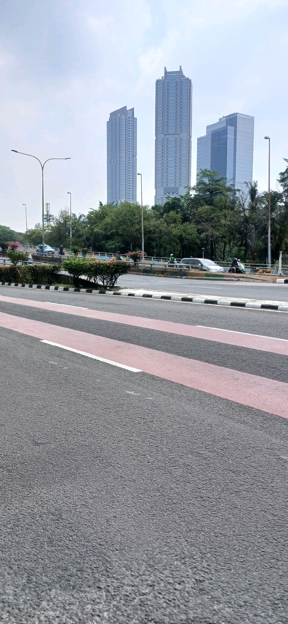 The image depicts a street scene in North Jakarta, Indonesia, with prominent skyscrapers in the background. In the foreground, a pedestrian crossing with red and white stripes is visible on an asphalt road. Several vehicles, including cars and motorcycles, are present on the road. Trees and decorative bushes line the median and sides of the road. Streetlights are also scattered throughout the scene. The sky is overcast. The overall impression is of a busy urban environment with a blend of modern architecture and greenery.