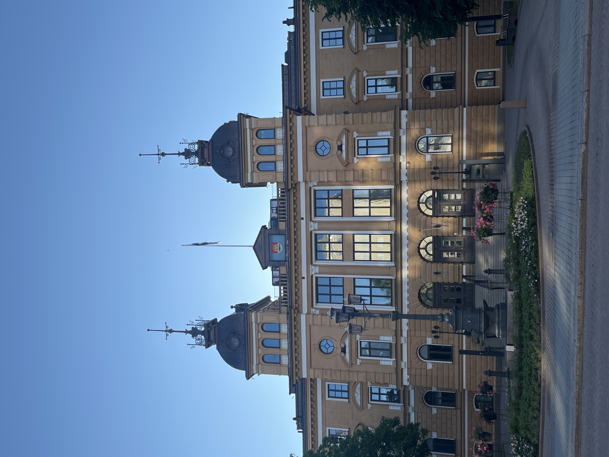 The image shows the Oulu City Hall, a prominent landmark in Oulu, Finland. The building is made of light-colored stone and features a symmetrical facade with two prominent towers topped with decorative spires. The main entrance is accessed by a grand staircase, flanked by ornate lampposts. Several windows, some arched and some rectangular, are visible on the facade, reflecting the clear blue sky. Flower boxes with pink and white flowers are placed near the entrance and along the surrounding pathways. The foreground shows a paved area with a circular garden in the center, also adorned with flowers and greenery. The sky is a deep blue, indicating it is daytime, likely late afternoon or early evening, given the soft lighting. There are no people visible in the image.