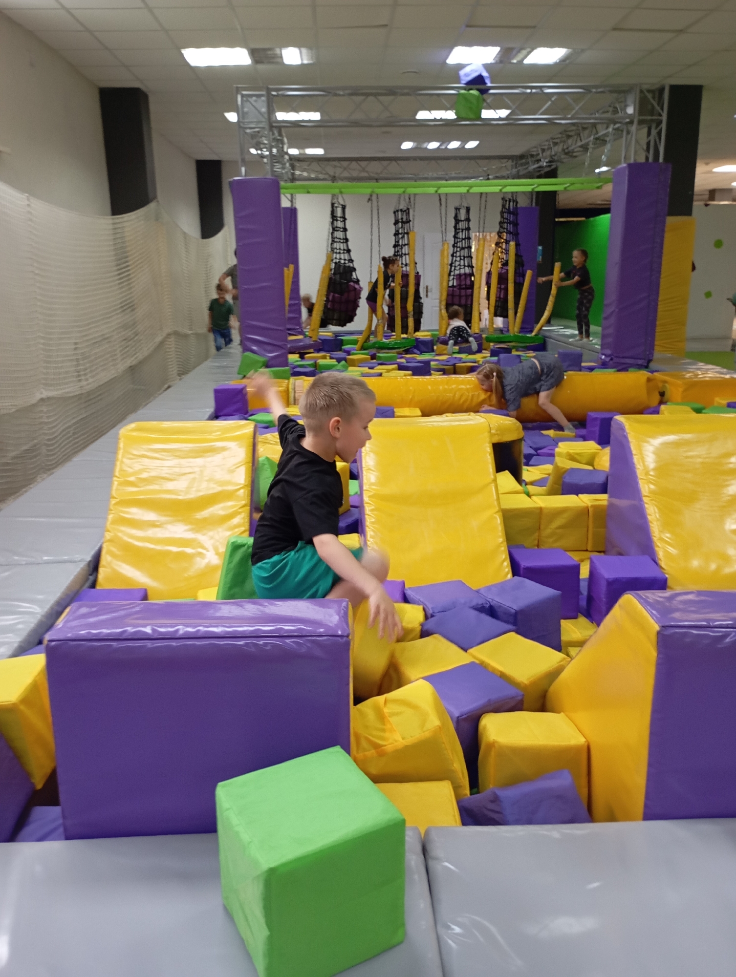 The image shows a group of children in an indoor play area. A boy wearing a black shirt and green shorts is in the foreground, seemingly jumping or climbing among yellow, purple, and green foam blocks. In the background, there are other children playing near hanging rope structures and padded columns, with a safety net visible on one side. The play area appears to be well-lit with artificial lighting. The city and state are unknown.
