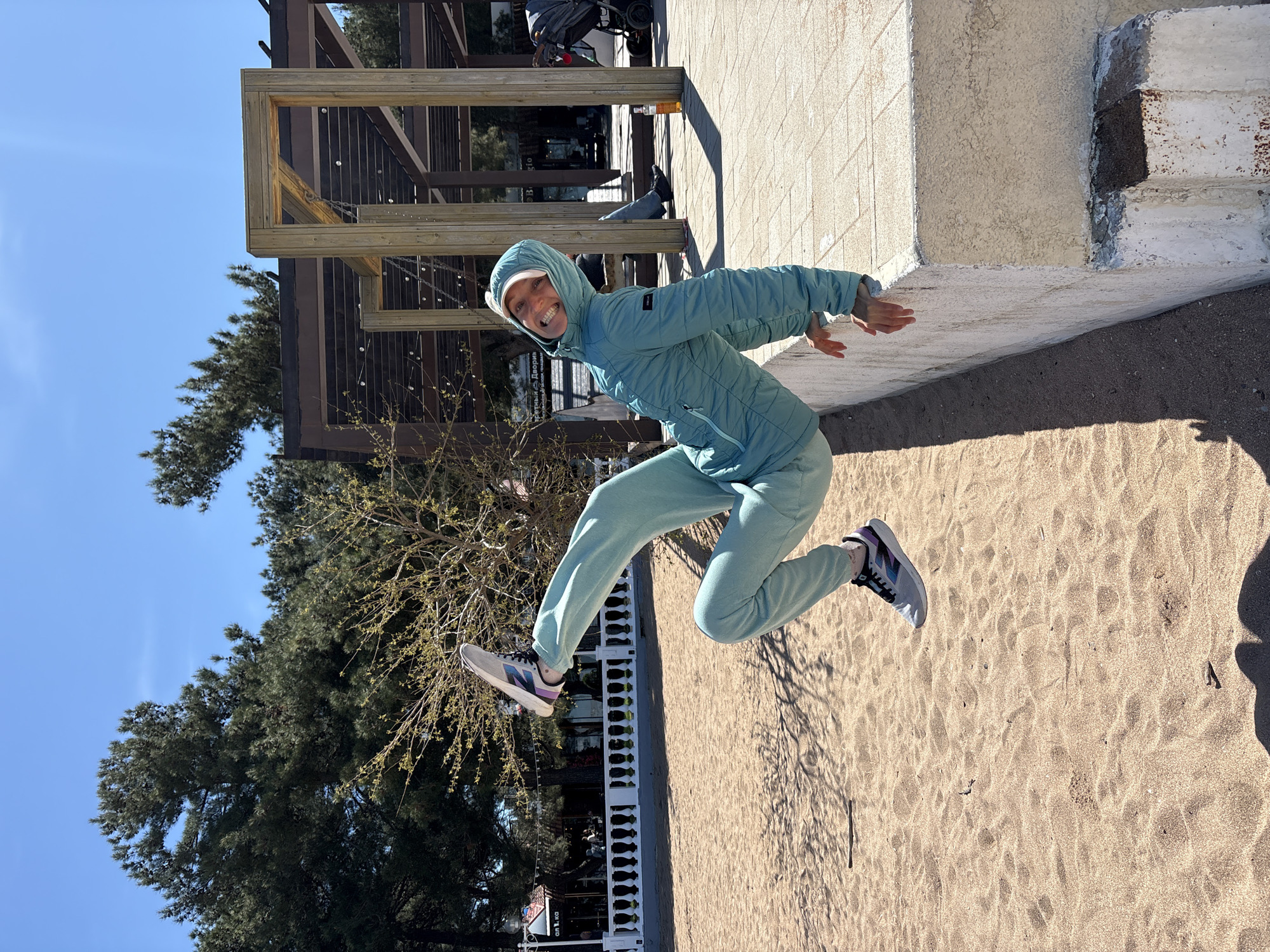 An energetic young woman is captured mid-air, joyful and smiling broadly, against a clear blue sky. She is dressed in a matching light blue or mint green hooded puffer jacket and sweatpants, paired with grey sneakers featuring purple and black accents. Her arms are slightly extended, and her legs are bent as she leaps, creating a dynamic pose.
The setting appears to be an outdoor recreational area, likely a sandy beach or park space, with sandy ground visible in the foreground and beneath her. To her right, a concrete wall or ledge runs alongside the sandy area, leading to a paved surface beyond. In the background, there are various wooden structures, possibly pergolas or parts of an outdoor installation, casting distinct shadows. Mature green trees, resembling pines, line the background, adding to the natural environment.
The bright sunlight suggests a clear, sunny day, with long shadows visible on the sand, indicating it's likely morning or afternoon. Her expression conveys happiness and freedom. In the deeper background, partially obscured, a sign with Cyrillic text can be seen, reading "Третий ДВОР" (Tretiy DVOR), which translates to "Third Yard" or "Third Court," possibly indicating a local establishment like a cafe or recreational zone in Gelendzhik, Russia. A stroller is also faintly visible further back.