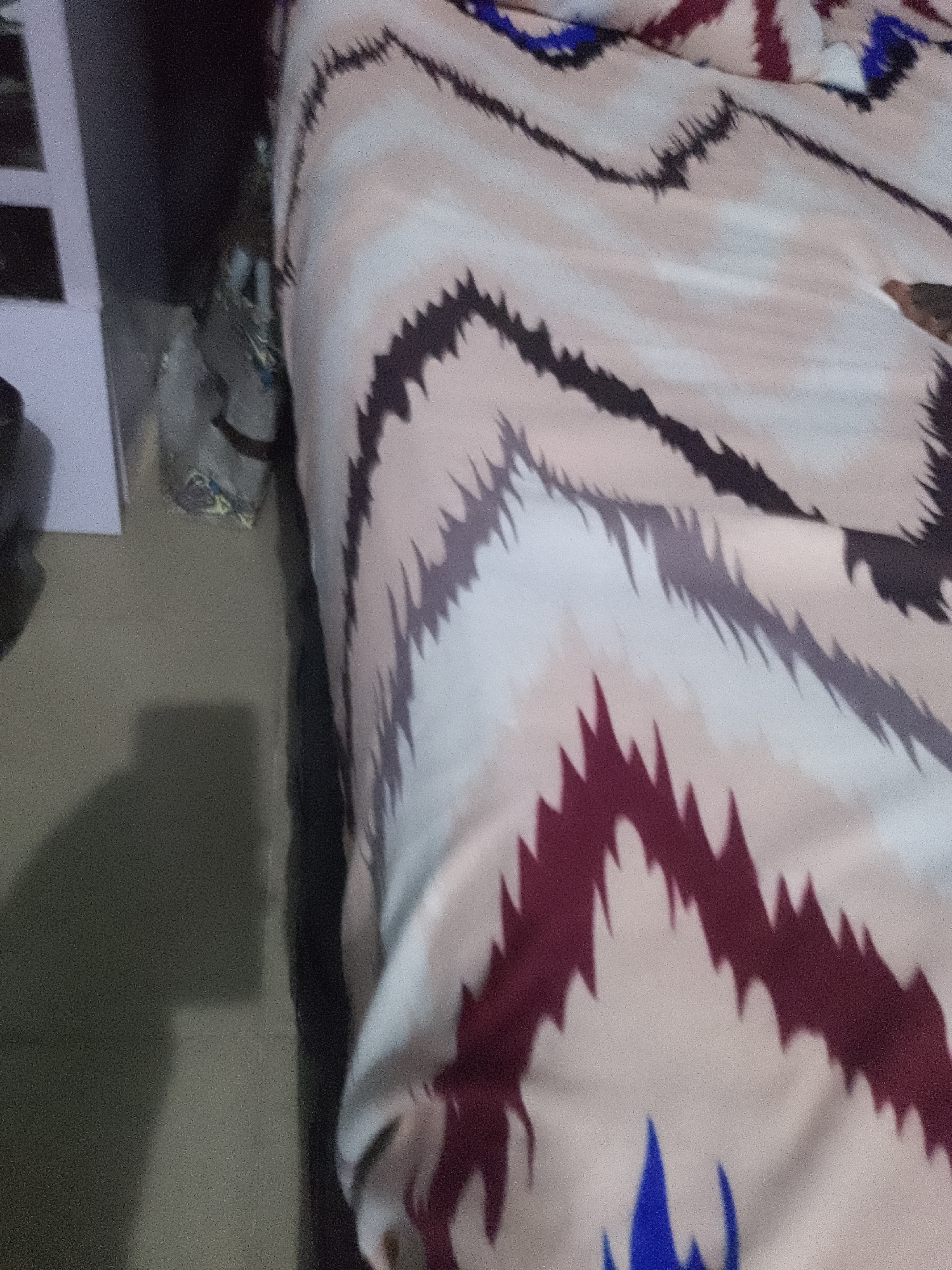 A close-up, angled shot shows a rumpled bedsheet with a geometric pattern. The pattern features jagged, zigzagging bands of dark purple, gray, and light tan against a pale pinkish background. Hints of blue appear in some of the peaks of the pattern. A dark, indistinct shadow is cast on the floor to the left of the bedsheet, and a white vertical object, possibly part of a piece of furniture, is partially visible on the far left. The lighting suggests an indoor environment.