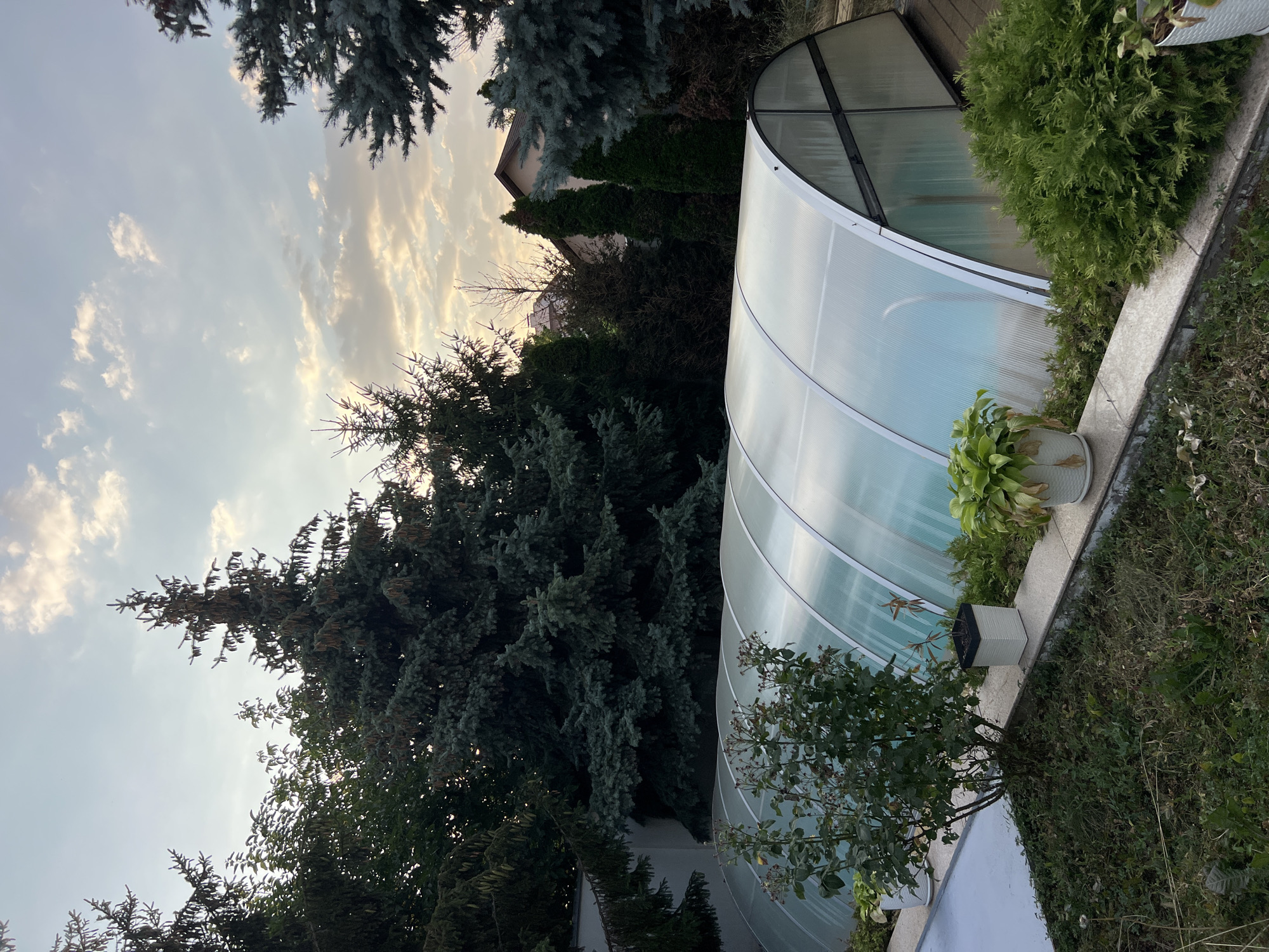 The image depicts an outdoor scene featuring a covered swimming pool in what appears to be a residential backyard. The pool enclosure is made of translucent, ribbed material supported by a white metal frame, creating a tunnel-like structure over the pool. The water within the pool is visible through the translucent covering and appears to be a light blue color. Surrounding the pool are various types of evergreen trees and shrubs, including tall fir trees and a denser, bush-like evergreen in the foreground. There are also some potted plants placed on a low, stone wall that runs along the edge of the pool enclosure. In the background, parts of residential houses with pitched roofs are visible, nestled among the trees. The sky above is partly cloudy, with patches of sunlight breaking through, suggesting it is either sunrise or sunset. The overall impression is a well-maintained outdoor space, likely a private residence. The location is described as the city of Fontanka, Ukraine. There are no people or animals visible in the image.