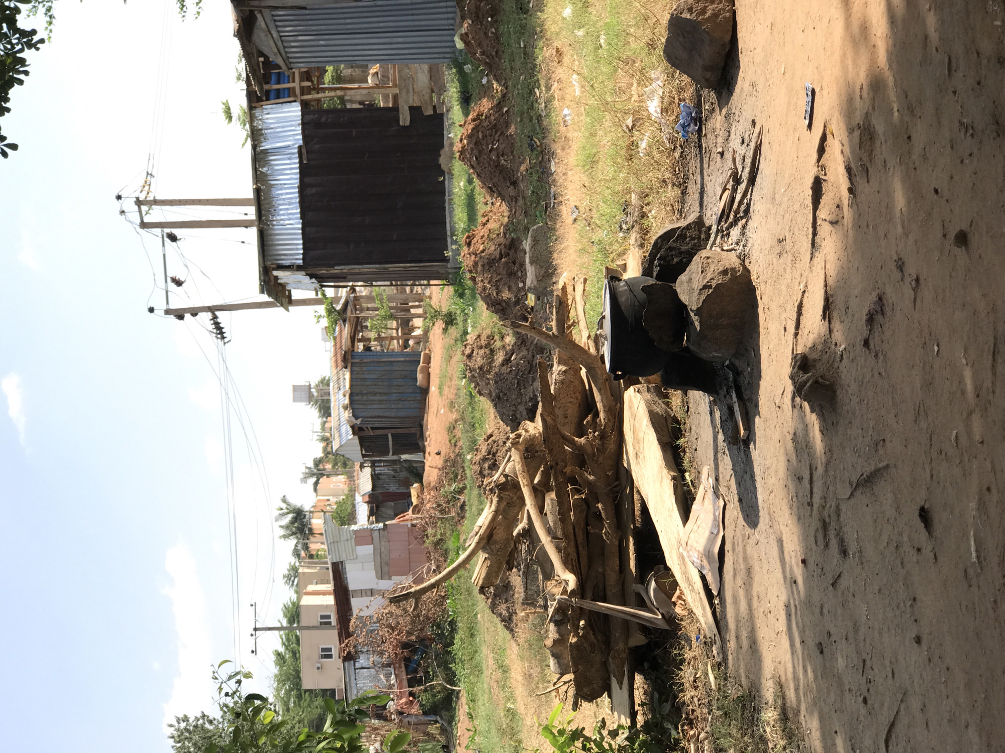The image depicts an outdoor scene, potentially in a less developed area in the city of Abuja, Nigeria. The foreground is dominated by a pile of firewood and large rocks, near what appears to be a small cooking area with blackened stones and a pot. The ground is bare earth, dusty and showing signs of recent use. In the background, there are several corrugated metal structures, likely shacks or small shops. Further back, there are more permanent buildings, including what appears to be residential apartments. Utility poles with power lines are also visible. The environment is a mix of dry vegetation, grass, and bare ground. There is no evidence of human presence.