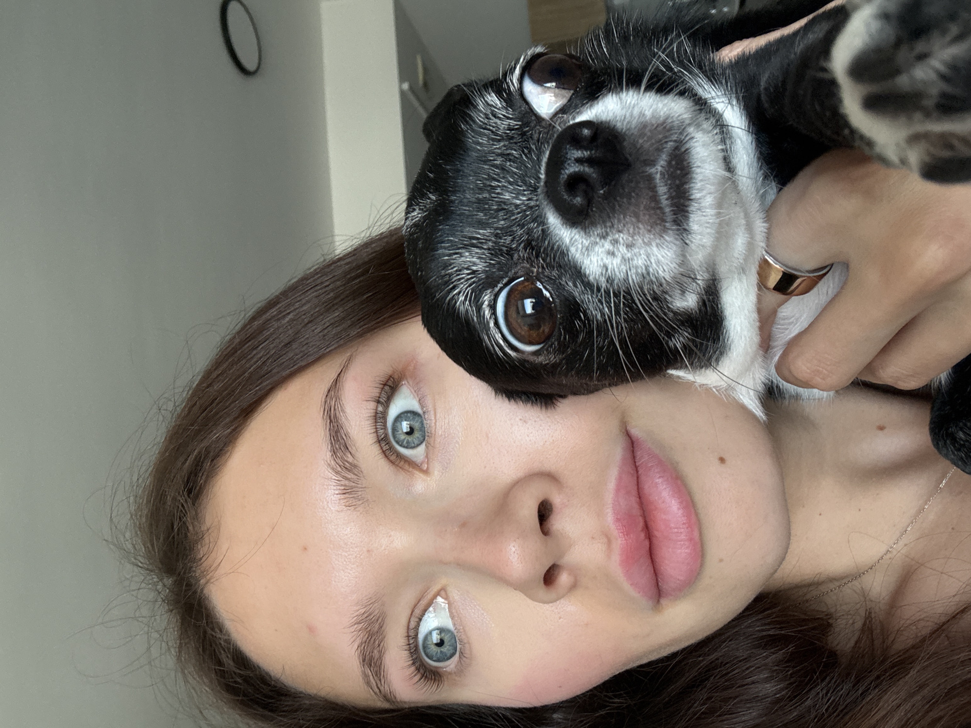 A close-up, portrait-oriented image shows a young woman and a small dog looking directly at the camera. The image, originally rotated 90 degrees, features the woman on the left and the dog held up beside her face on the right.
The woman has fair skin, prominent blue eyes, and long dark brown hair that frames her face. Her lips are a natural light pink, and she wears a delicate gold chain necklace. Her left hand is visible on the right side of the frame, holding the dog, and she wears a two-toned (gold and silver) ring on her ring finger.
The dog is small, with black fur and distinct white markings around its muzzle, chin, and chest, as well as some graying around its eyes. It has large, dark brown eyes that are wide and expressive, also looking directly into the camera. One of the dog's paws is visible in the upper right corner, resting near the woman's shoulder.
The background is a simple, light gray wall, with a black circular object, possibly a clock or mirror, visible in the upper left corner. The lighting is soft and even, highlighting their features. Both subjects appear calm, with the woman showing a slight smile and the dog an alert expression. There is no visible text in the image.
