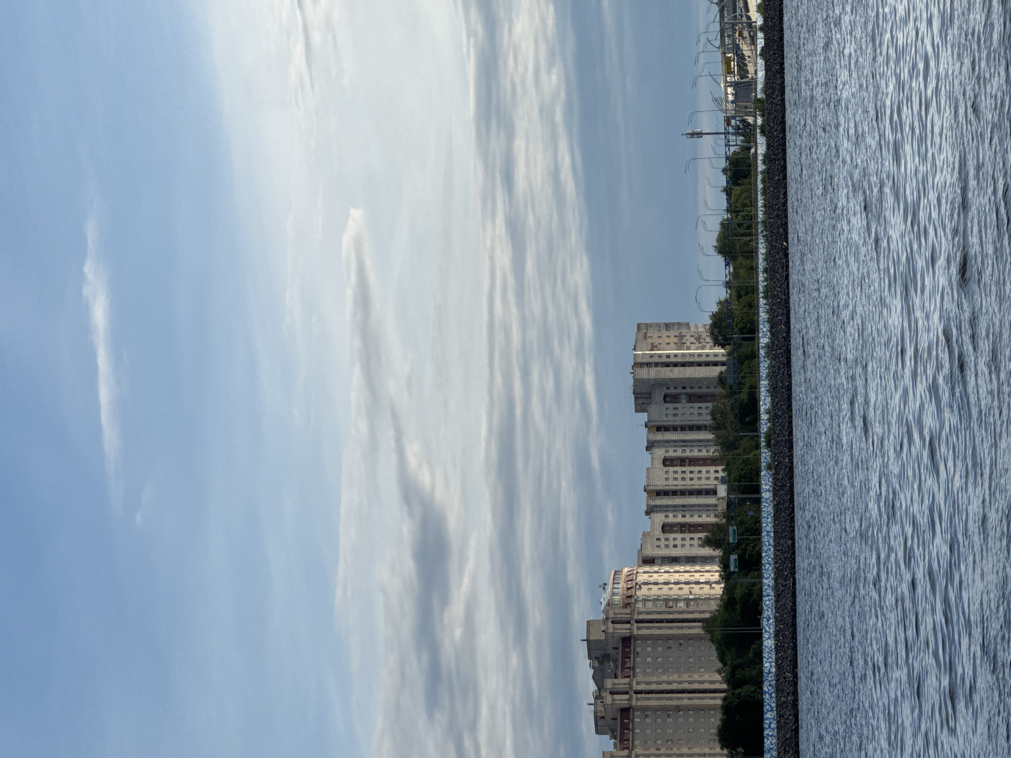 This image depicts a section of Saint Petersburg, Russia, along a body of water. The foreground is dominated by a stretch of rippling blue water. Behind the water, a low, dark stone embankment with a patterned blue trim separates the water from the land. Beyond the embankment, a line of trees runs horizontally across the mid-ground. Rising behind the trees are several tall, multi-story apartment buildings, characteristic of Soviet-era architecture, with many windows and balconies. In the background, there are visible elements of urban infrastructure, including streetlights, a telecommunication tower, and what appears to be a bridge or overpass system. The sky above is a mix of blue sky and soft, scattered clouds. The overall scene suggests a residential area adjacent to a waterfront, possibly a canal or bay, within the city.