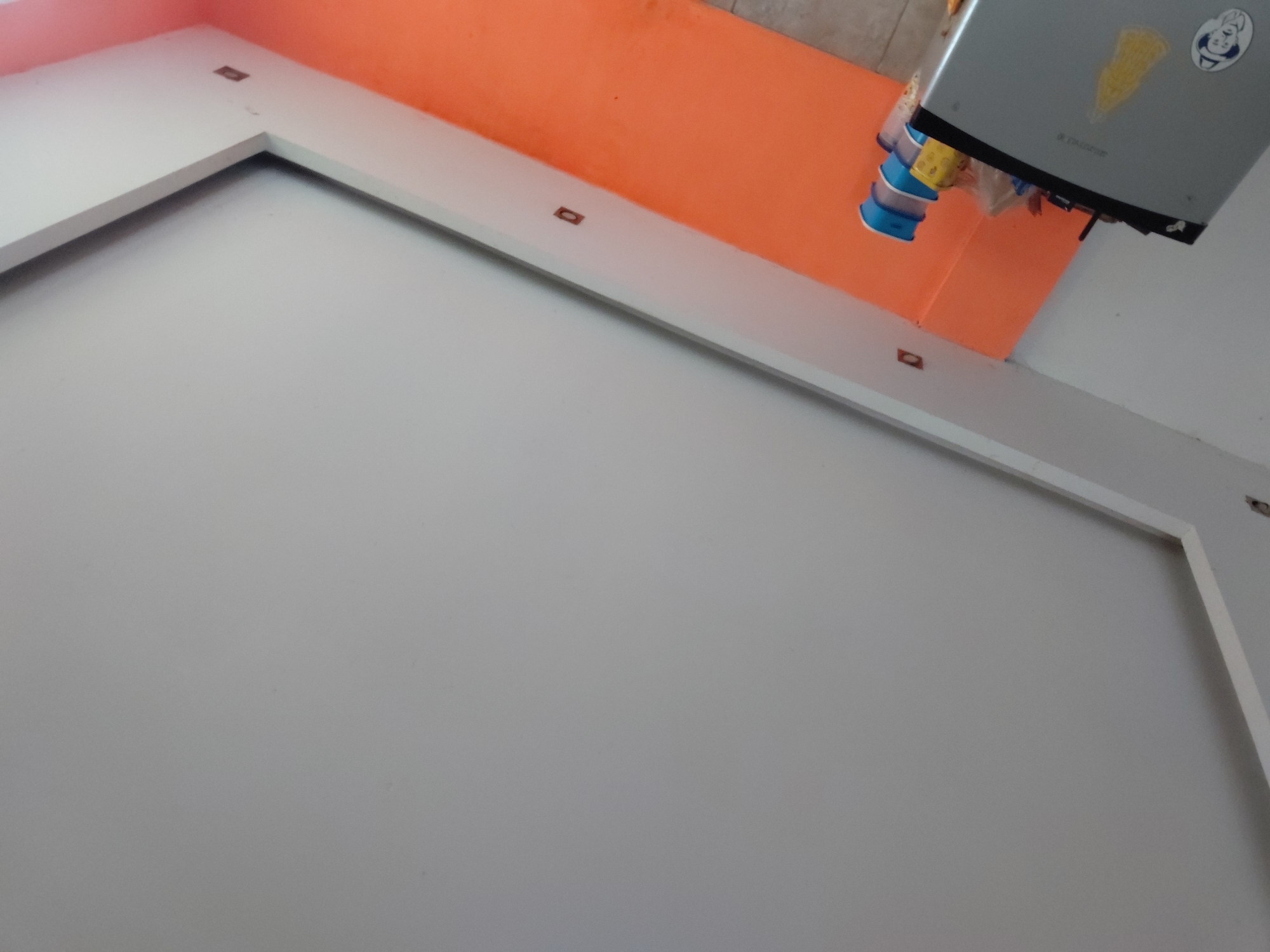 The image captures an indoor scene from a low, tilted perspective, looking upwards towards a corner. A prominent orange wall is visible in the upper section, meeting a lighter-colored wall or ceiling. A wide, white trim or countertop runs along the base of the orange wall, extending across the image.
Several small, dark, rectangular objects, likely electrical outlets or mounting points, are embedded at intervals within the white trim. In the upper right corner, a silver-grey microwave oven is partially visible. It features a yellow, triangular sticker and a circular white sticker depicting a cartoon character, possibly a chef, with some illegible text. The brand "DURABRAND" is faintly visible on the microwave. Stacked blue plastic containers or cups are present underneath the appliance. The foreground and the majority of the left side and bottom of the image consist of a plain, light grey or off-white wall or ceiling surface. No individuals are present, and no activity is taking place. The lighting suggests an indoor environment.