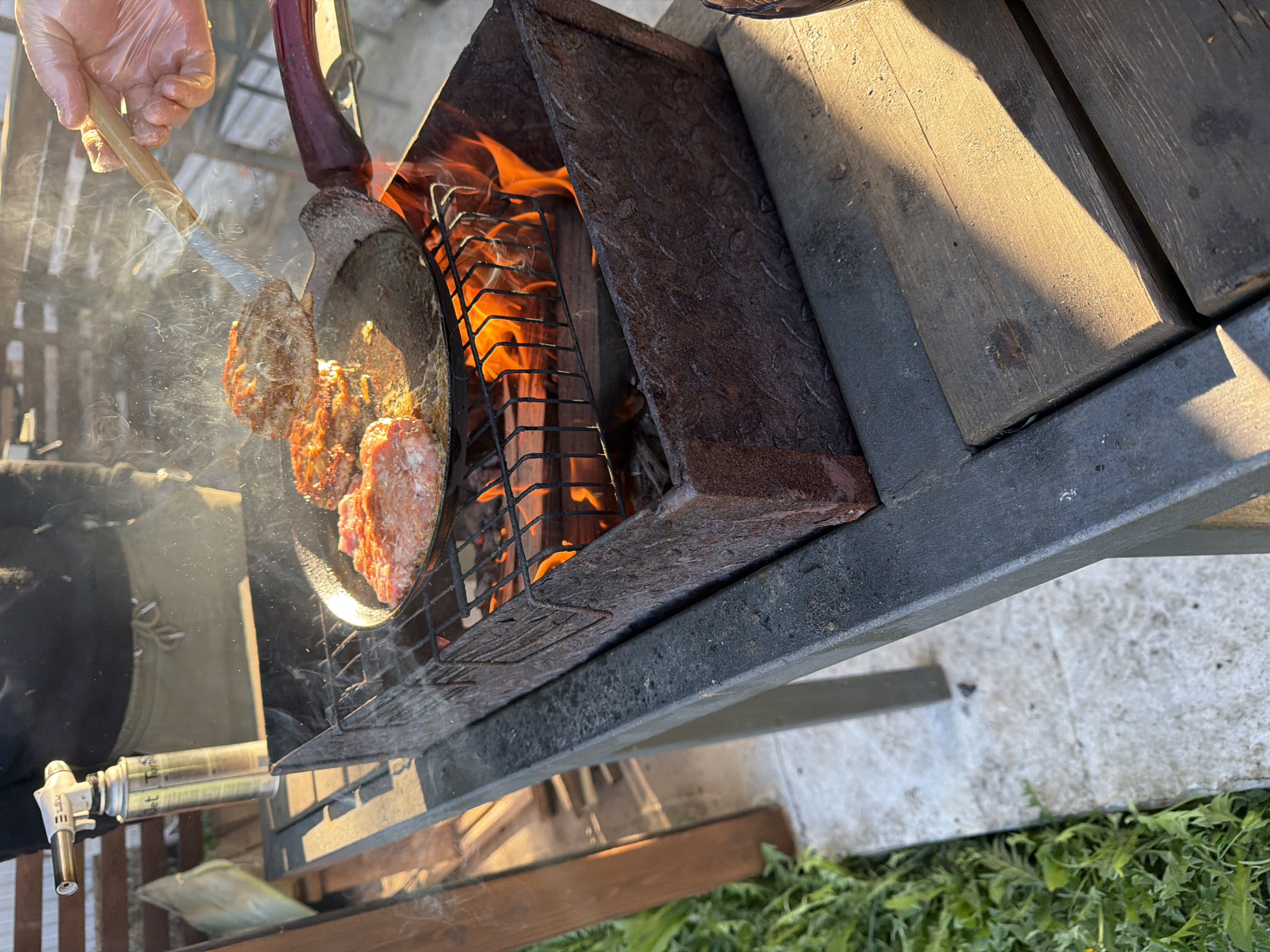 The image shows a cooking scene, possibly outdoors in Большая Шильна, Russia. Someone wearing a clear plastic glove is using tongs to lift a cooked patty from a cast iron pan. The pan is sitting on a grill over a fire inside a metal structure that appears to be part of an outdoor cooking station. The grill has a metal hood above the fire, and a wooden tabletop extends to the right. In the background, a gas torch is visible. The presence of grass suggests the scene is located in a yard or garden. Smoke is present.