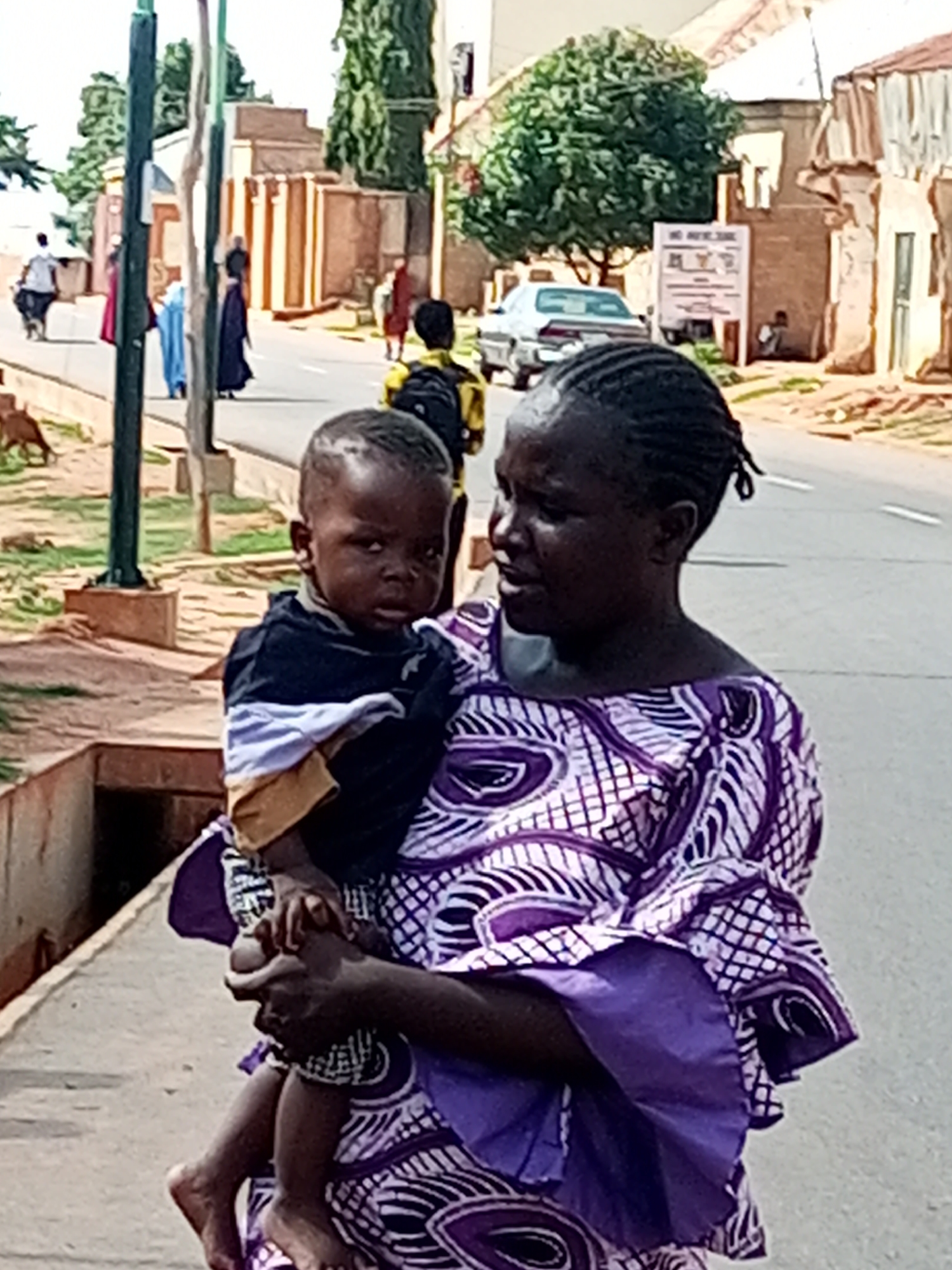 In Tempure, Nigeria, a dark-skinned woman carrying a young child on her hip walks along a paved sidewalk. The woman wears a purple and white patterned dress with a matching purple underskirt. The child is dressed in a dark blue shirt with horizontal white and tan stripes on the sleeves and chest. The background shows buildings constructed from red brick with arched windows and doorways, and a large green tree. A white car is parked on the street in the distance, along with several other individuals walking. A street lamp stands on the left side of the image.