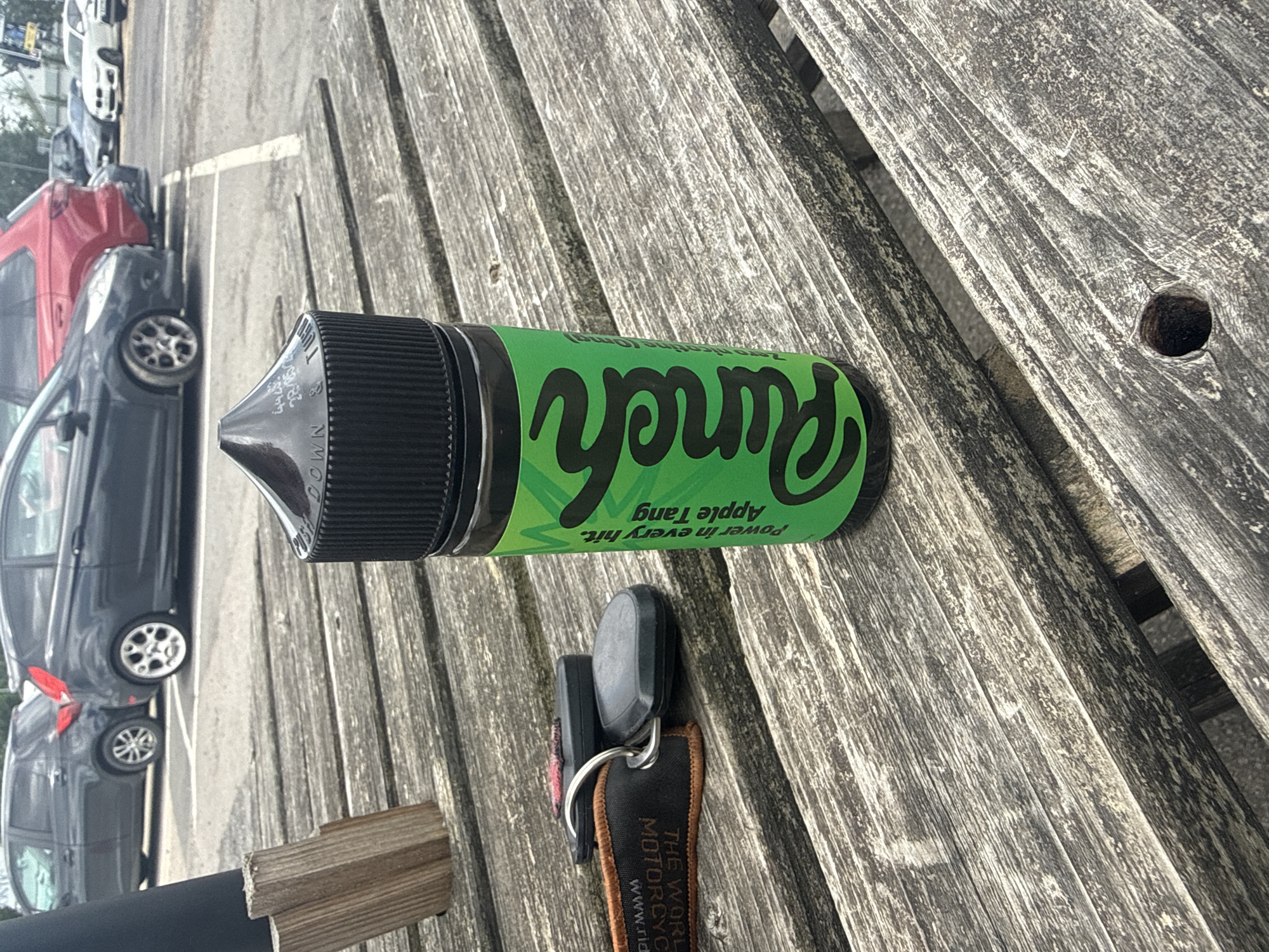 The image shows a bottle of e-liquid with a green label on a weathered wooden picnic table. The bottle has a black cap and writing in black on the cap. The label on the bottle is green with black lettering that says "Punch" in large stylized script, and "Power in every hit, Apple Tang" in smaller text below. To the left of the bottle are a set of car keys attached to a black and orange fob that reads "THE WORLD'S MOST CYCLIST". In the background, parked cars are visible, including a red Audi and dark grey Ford Fiesta. The scene appears to be an outdoor area, possibly a car park or rest stop. The location is identified as Tewkesbury, United Kingdom.