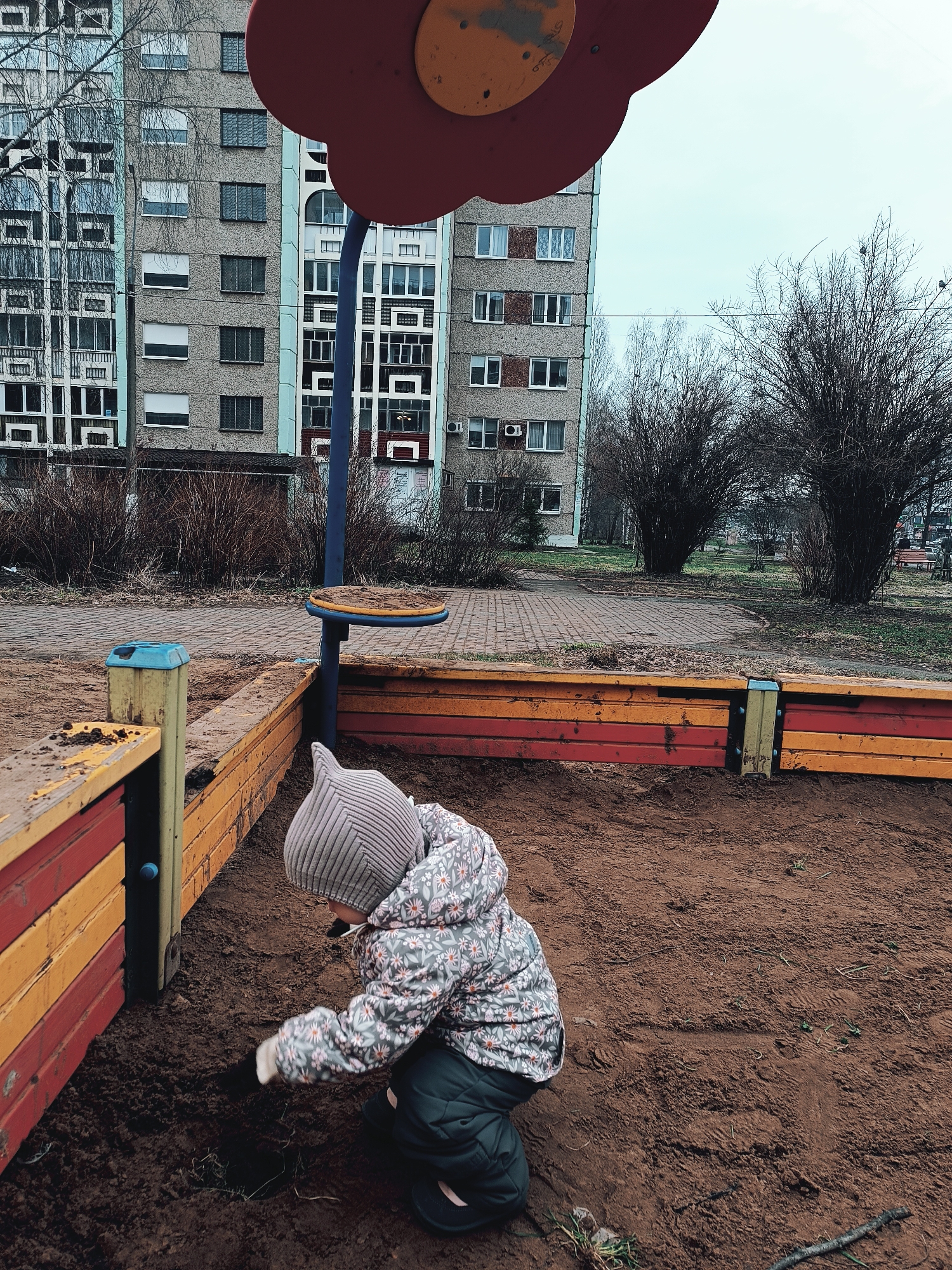 A child is digging in a sandbox in a playground in Izhevsk, Russia. The sandbox has wooden borders painted in yellow and red. A playground structure with a red flower shape overhead and a blue pole stands nearby. Behind the playground, multi-story apartment buildings dominate the background, along with bare trees. The child is wearing a patterned jacket, a gray striped hat, and dark pants. The sand in the sandbox is dark and slightly muddy. The scene is overcast.