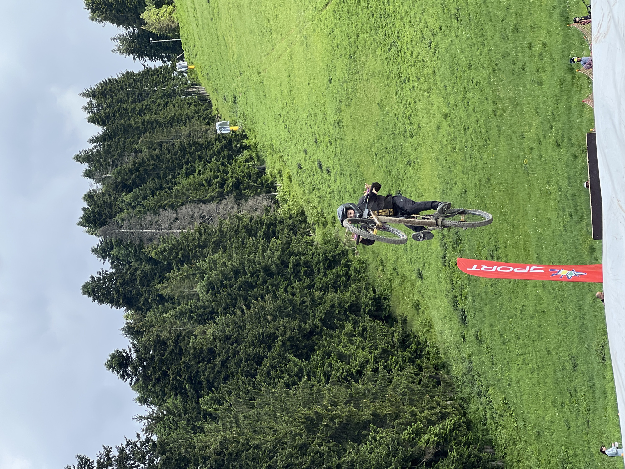 A mountain biker in mid-air performs a trick while riding a bicycle down a grassy slope. The biker is wearing a helmet and black clothing. A red banner with the word "SPORT" is visible in the foreground. Trees are visible in the background, and the sky is cloudy. The location is a ski resort or a similar outdoor recreational area.