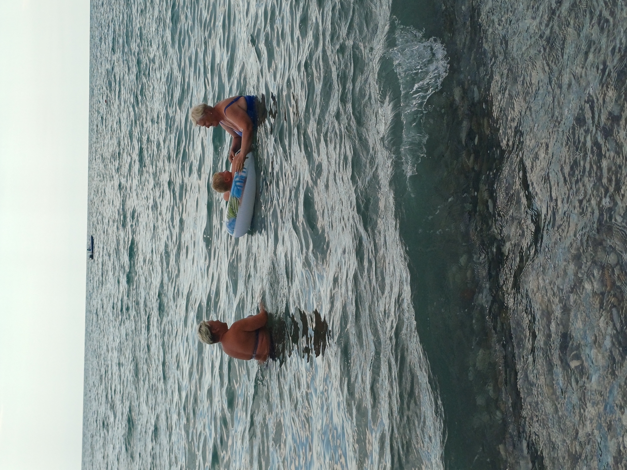 The image shows three people in the sea at Sochi, Russia. Two adults and a child are in the water. One adult, a woman with blonde hair, is wearing a blue swimsuit and is assisting a child who is in a yellow and blue inflatable ring. The child is also in the water. Another adult, also a woman with blonde hair, is in the water facing away from the camera. The sea is choppy, with visible waves. The sun appears to be out, as indicated by the bright sky on the left side of the image and the reflections on the water. The water is clear enough to see the rocky bottom in some areas. The scene appears to be a recreational outing, likely at a beach or resort.