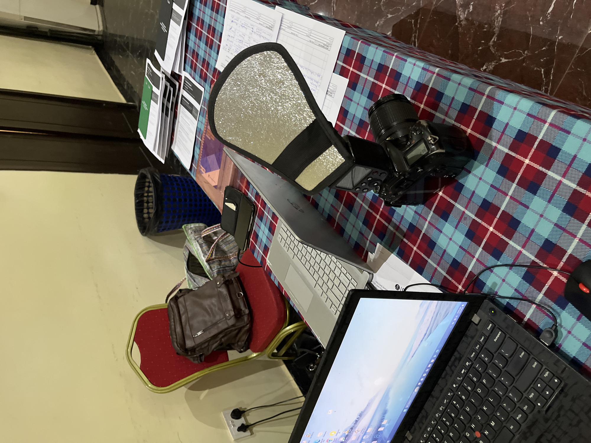 The image shows a cluttered workspace on a table covered with a red and blue plaid tablecloth. Several objects are present, including two laptops, a camera with a flash diffuser, various documents, a handbag, and a blue patterned basket. One laptop, with a black keyboard, is closer to the viewer and shows a brightly lit screen. Another, with a silver keyboard, lies further back on the table. The camera is positioned near the edge of the table on the right side, along with the flash diffuser that is attached to it. The handbag sits on a small red chair. A power strip is visible near the bottom left corner of the image, suggesting an indoor setting where electronic devices are being used. The background is blurred, but a portion of a wall can be seen, contributing to the impression of an indoor location. The location is unknown.
