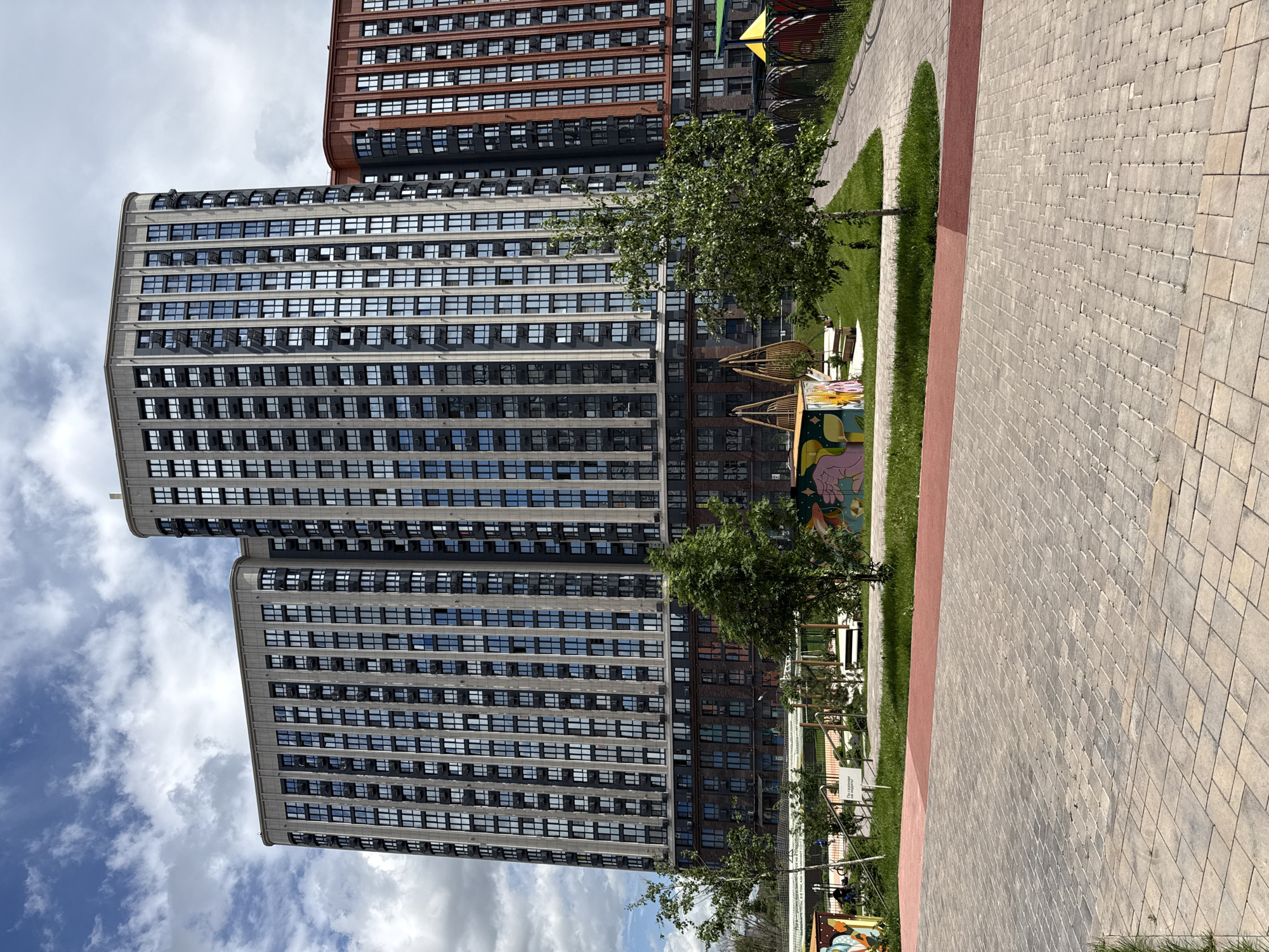 The image shows a high-rise building complex in what is likely a residential area. The buildings are modern in design, featuring numerous windows and varying colors, with one tower being primarily gray and another showing red brick elements. Between the towers, there's a glimpse of a blue sky dotted with clouds.
In the foreground is a paved pathway bordered by a strip of green grass. A children's playground is situated near the walkway, with colorful play structures and swings. Trees are sparsely planted in the grass area.
Based on the provided information, the location is in the city of Ovoshchnoy, Russia. However, without further details, no specific people, actions, or interactions are visible.