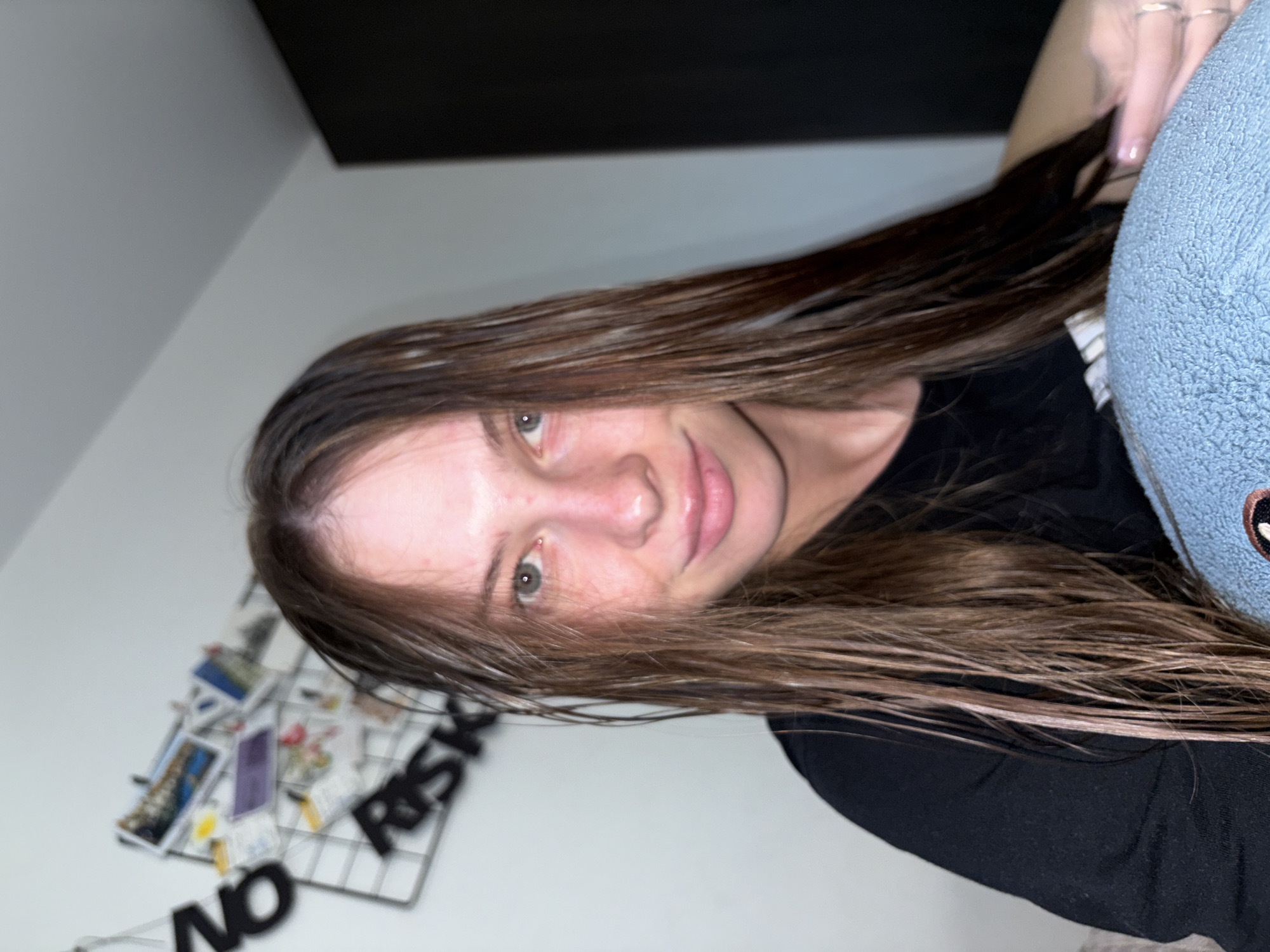A young woman with wet, brown hair is looking at the camera. She has fair skin and light-colored eyes, and is wearing a black t-shirt. In the background, there is a white wall with a grid decorated with postcards and black lettering that appears to spell out "NO RISK". The woman is holding a blue, textured object in her lap. The image appears to have been taken indoors, in a residential setting. There are no specific visual indicators that place the location in Perm, Russia.