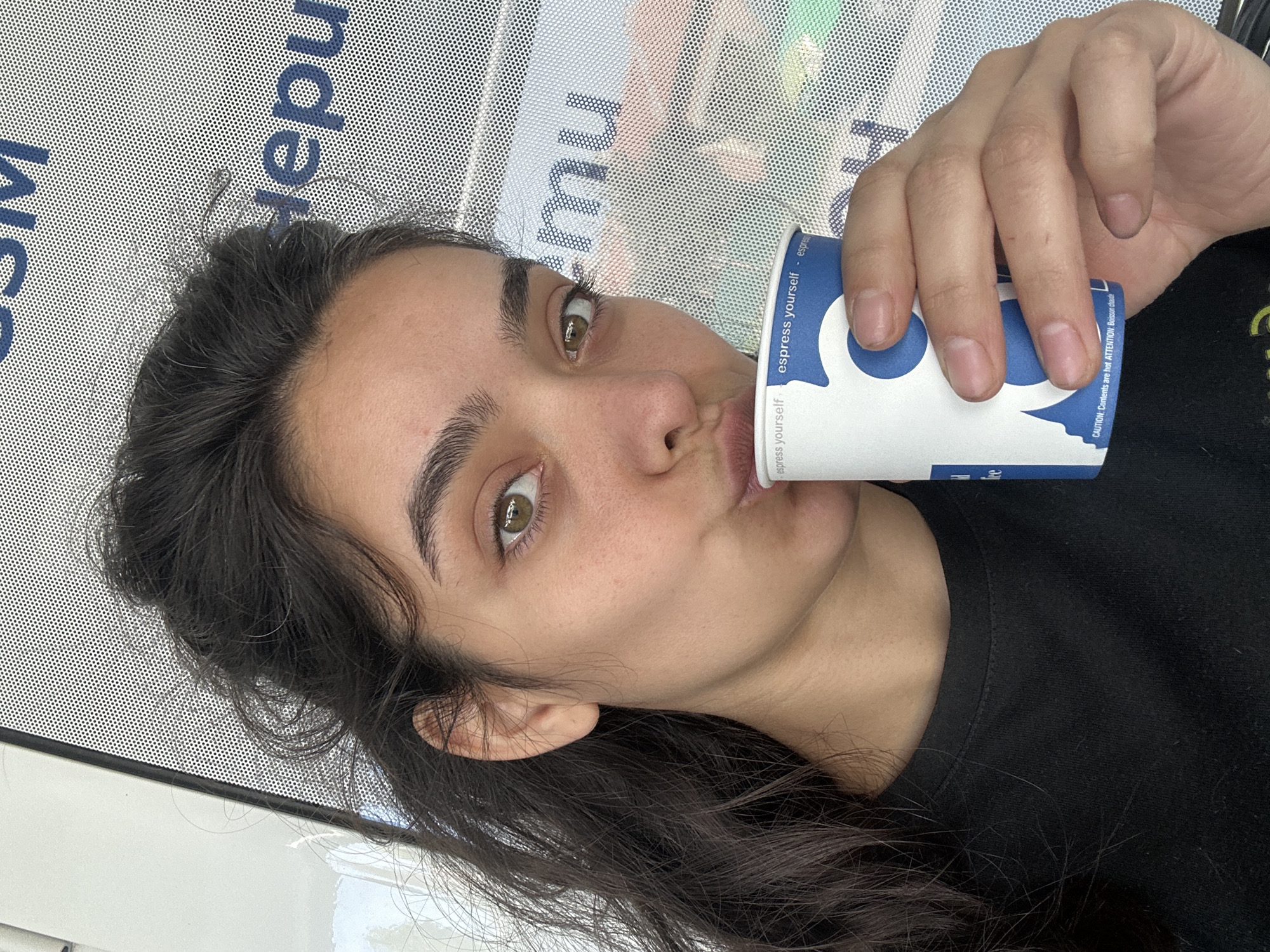 A close-up, waist-up shot captures a young woman with dark, wavy hair and light green eyes, leaning forward and puckering her lips to sip from a blue and white paper coffee cup. She is wearing a black t-shirt. Her right hand holds the cup. The background is a textured white surface with a pattern of small dots, behind which blurred green and pink shapes are visible, suggesting an outdoor or semi-outdoor environment. Text on the cup reads "espress yourself." The woman's expression is playful, with her eyebrows raised and eyes looking directly at the camera. The scene appears to be outdoors, possibly in a public space. There is no indication of the location being Malevo, Bulgaria.