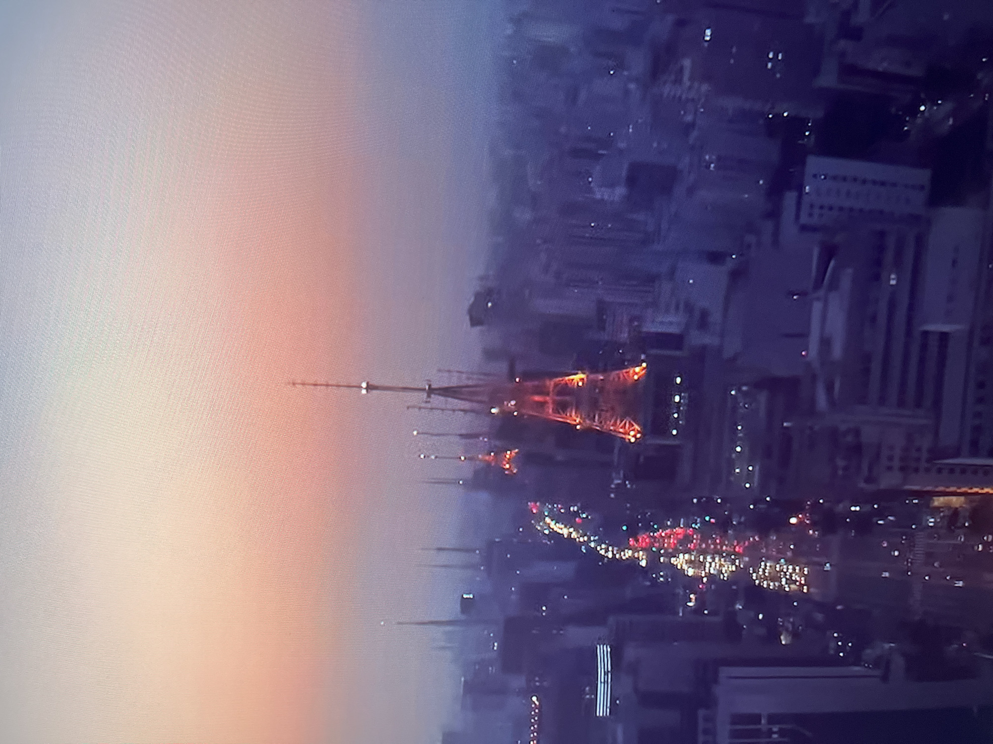 An aerial, wide-angle shot captures a city skyline at dusk. The sky transitions from a soft orange and pink near the horizon to a pale blue and white overhead. Several tall, latticed towers, some illuminated with red and orange lights, dominate the view, piercing the hazy atmosphere. Buildings of various heights, appearing as dark silhouettes with scattered lights, form a dense urban landscape. A busy street, illuminated by the headlights and taillights of numerous vehicles, snakes through the city, creating streaks of red and white light. The overall impression is one of a vibrant, bustling metropolis under a serene, fading sky.