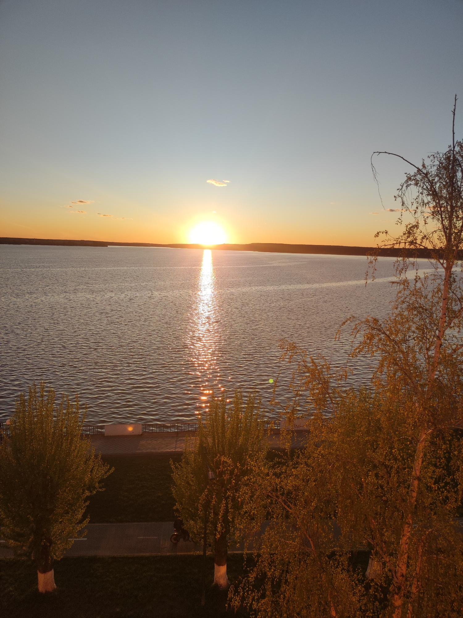 The image captures a sunset over a body of water in Votkinsk, Russia. The sun is setting directly in the center of the frame, reflecting a bright line of light across the water's surface. The sky transitions from a pale blue to a warm orange hue near the horizon. The water appears calm with slight ripples.
In the foreground, trees with green and golden foliage line the shoreline. A paved promenade with a railing is visible between the trees and the water. A street runs behind the promenade, and a cyclist rides on the street. The image suggests an urban landscape merging with a natural waterfront.