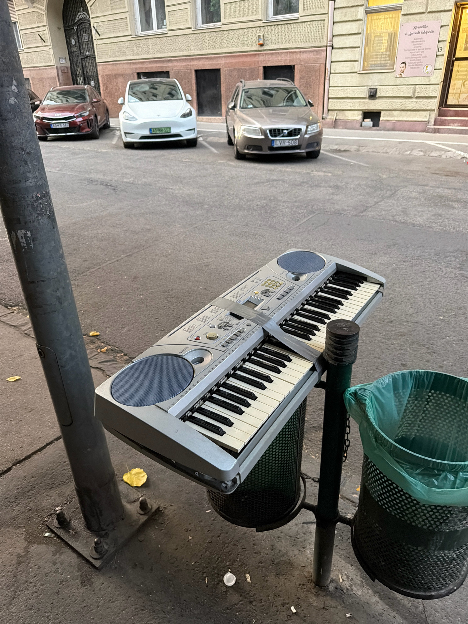 The image depicts a street scene in Budapest, Hungary, focusing on an electronic keyboard propped up on a trash can. The keyboard is silver with black and white keys, and appears to be somewhat damaged, with tape visible holding parts together. It is positioned on top of a black metal trash can, which has a green plastic liner. Next to this trash can is another identical one, also with a green liner. The keyboard and trash cans are situated next to a grey metal lamppost. The ground is asphalt, with some fallen yellow leaves scattered around. In the background, there are parked cars, including a white Tesla and a dark red Kia. Beyond the cars, there is a building with a light yellow facade and dark brown accents around the windows and doors. A poster with text and an image of a woman's face is visible on the building's facade. The lighting suggests it is daytime, with shadows cast on the ground.