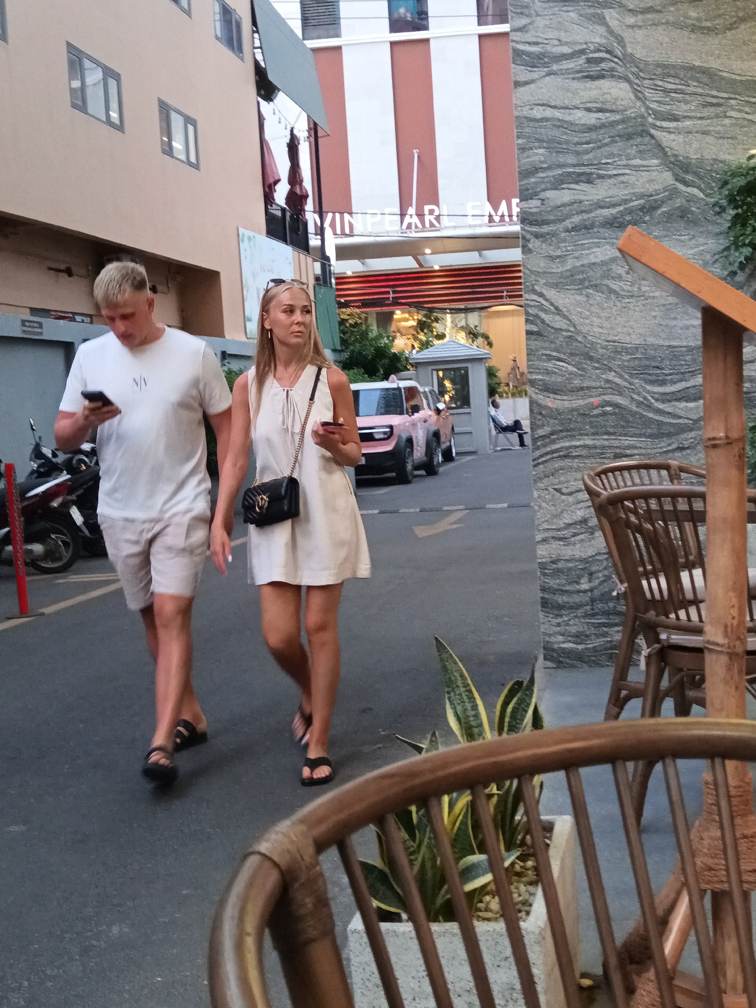 The image depicts a street scene in Nha Trang, Vietnam. In the foreground, a man and a woman are walking side-by-side. The man, wearing a white t-shirt and light-colored shorts, is looking down at his phone. The woman, dressed in a white dress, carries a black handbag and also holds a phone in her right hand. They are walking on an asphalt surface, possibly a sidewalk or pedestrian area. Behind them, a pink car is parked. The background features a building with signage that reads "PEARL EM". The scene appears to be an outdoor setting during daylight.