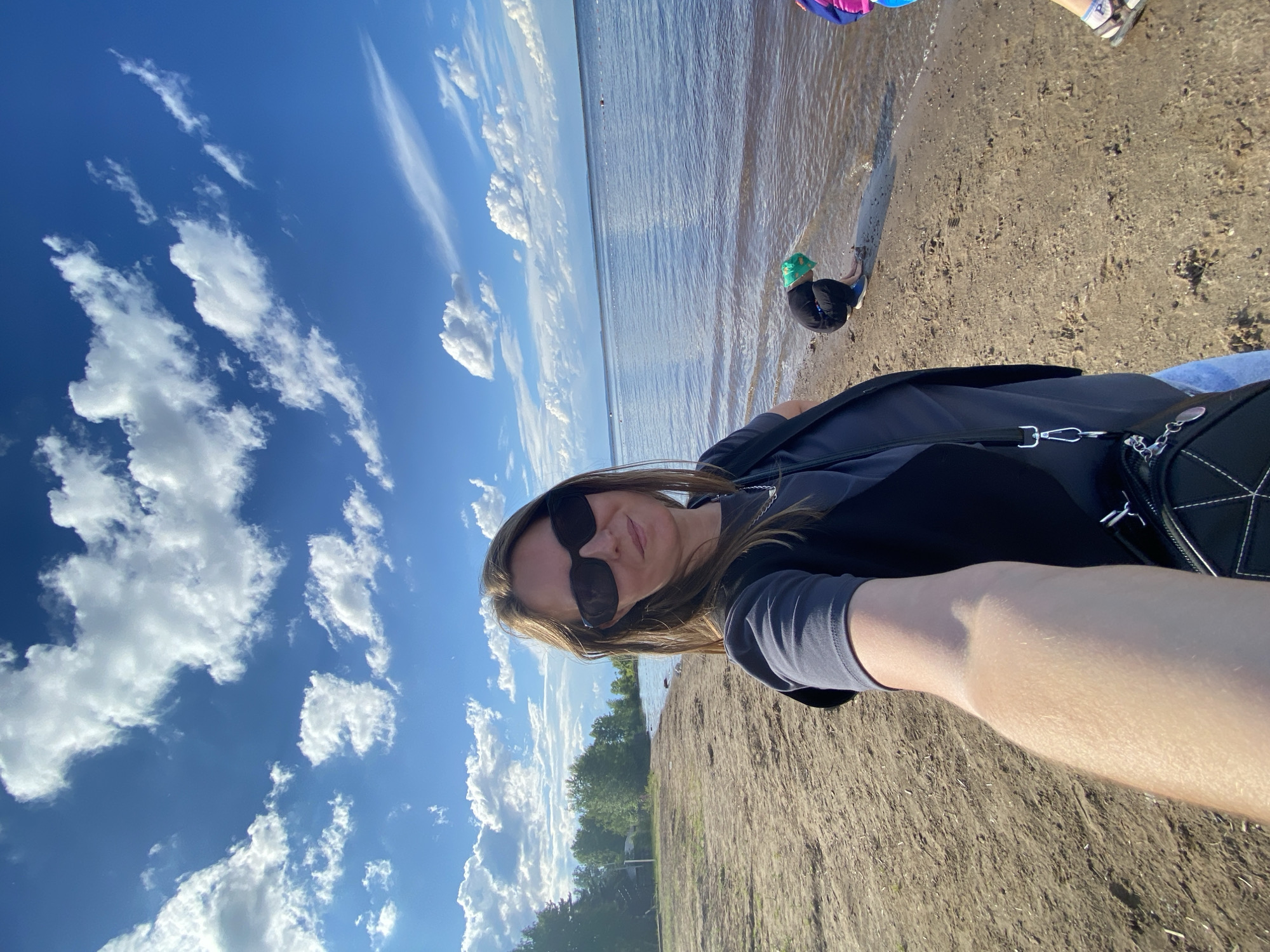 This is a selfie taken outdoors on a bright, sunny day with a clear blue sky filled with fluffy white clouds. The scene is a sandy beach bordering a body of water, likely a lake or river, with trees visible in the distance.
The main subject is a woman wearing sunglasses and a dark top, smiling at the camera. She is positioned in the foreground, with her arm extended to take the photo. In the background, a young child wearing a green hat is playing near the water's edge. Other people's legs and feet are visible at the top of the frame, suggesting a group outing.
The activity appears to be a casual day at the beach, with the woman capturing a moment of her family's outing. The sunlight indicates it is likely daytime, and the clear sky suggests fair weather. The presence of a beach and water points to a recreational location. No visible text is present in the image.