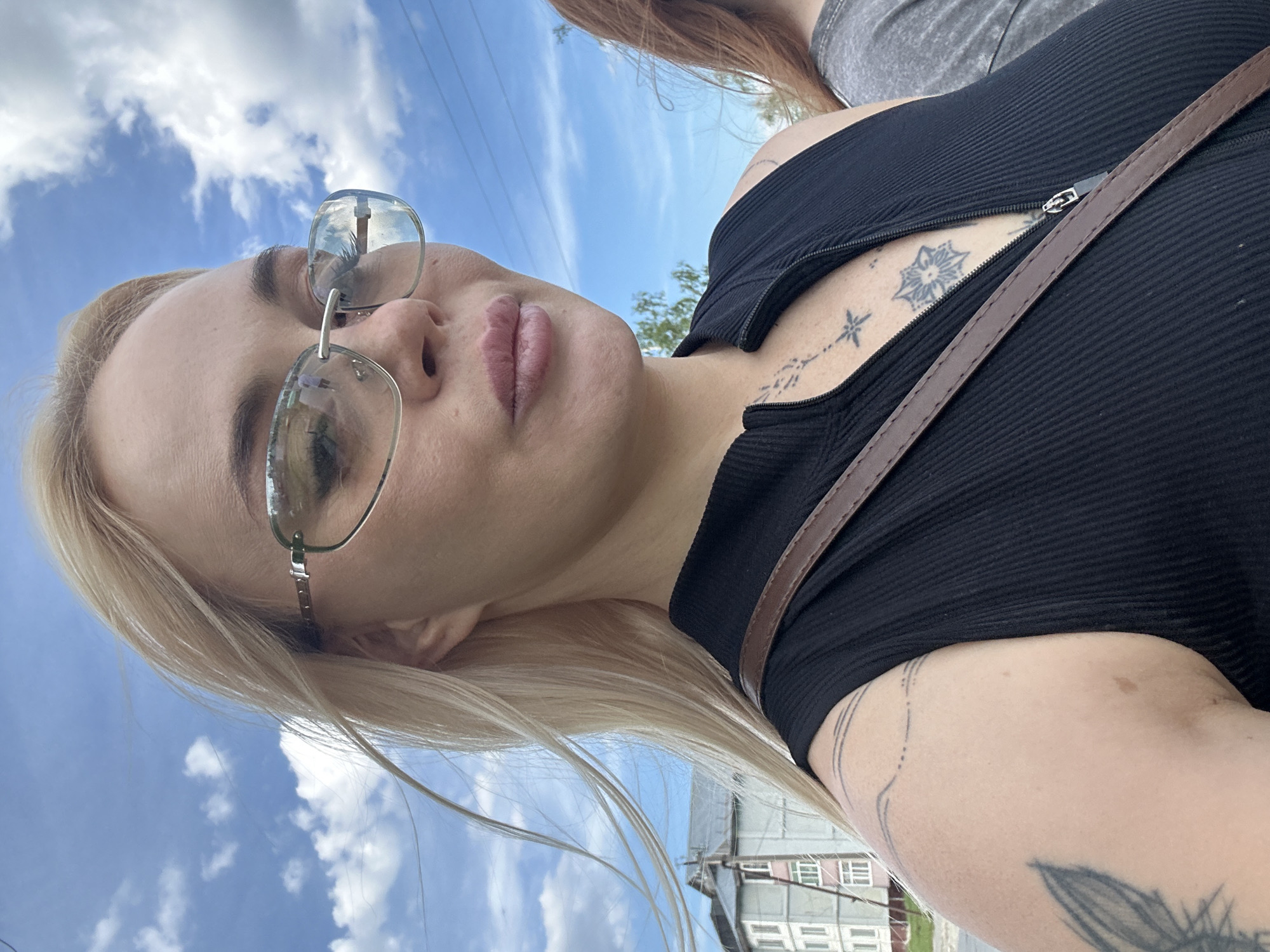 The image is a medium close-up, outdoor portrait of a blonde woman in an urban or semi-urban setting, taken from a slightly low angle. The woman, who is the primary subject, faces slightly to the right of the viewer with her head slightly tilted, and her gaze is directed generally towards the camera.
She has long, straight blonde hair, with some strands appearing to be gently blown by the wind. She wears rimless, lightly reflective sunglasses with silver-toned bridge and temple pieces, obscuring her eyes. Her lips are full and have a pink or nude-colored tint, appearing slightly parted or relaxed.
She is wearing a black, ribbed, sleeveless top with a high neckline and a short silver-toned zipper down the front, which is partially unzipped, revealing some cleavage. A brown leather or faux-leather strap from a bag is visible diagonally across her upper chest and over her right shoulder.
Several tattoos are visible on her skin. On her left upper arm and shoulder, there are multiple thin, curving lines, some solid and some dotted. On her chest, in the exposed area, there is a small star-like symbol just below the zipper, and further down, a larger, multi-pointed geometric or mandala-like design. Another thin vertical line with a small star or cross-like symbol is visible higher on her chest.
To the right of the primary subject, the left side of another person's head and shoulder are visible. This person has reddish-brown hair and is wearing a gray, textured (possibly heathered) t-shirt.
The background consists of a bright blue sky with scattered white cumulus clouds. On the left side of the frame, behind the primary subject, there is a multi-story light-colored residential building with several windows. Another building with a darker roof is partially visible behind it. On the right side of the frame, some green foliage (trees or bushes) is visible, and several thin, dark utility lines (power or communication cables) stretch horizontally across the upper right portion of the sky.
The scene takes place in the city of Unknown, Unknown.
