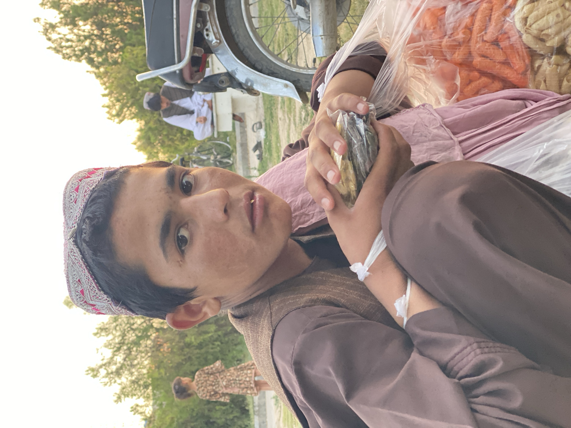 A young boy wearing a traditional embroidered cap and brown clothing is crouched down, holding a small package wrapped in plastic. He appears to be in an outdoor setting, possibly a market or roadside stall in Kandahar, Afghanistan, as indicated by the presence of other people and a motorcycle in the background, along with a display of produce, including what appear to be carrots. The sun is setting, casting a warm glow on the scene. A young girl in a patterned dress is visible in the background, and another person, also wearing traditional clothing, is seated behind a motorcycle. The overall atmosphere suggests a common, everyday scene in a bustling urban environment.