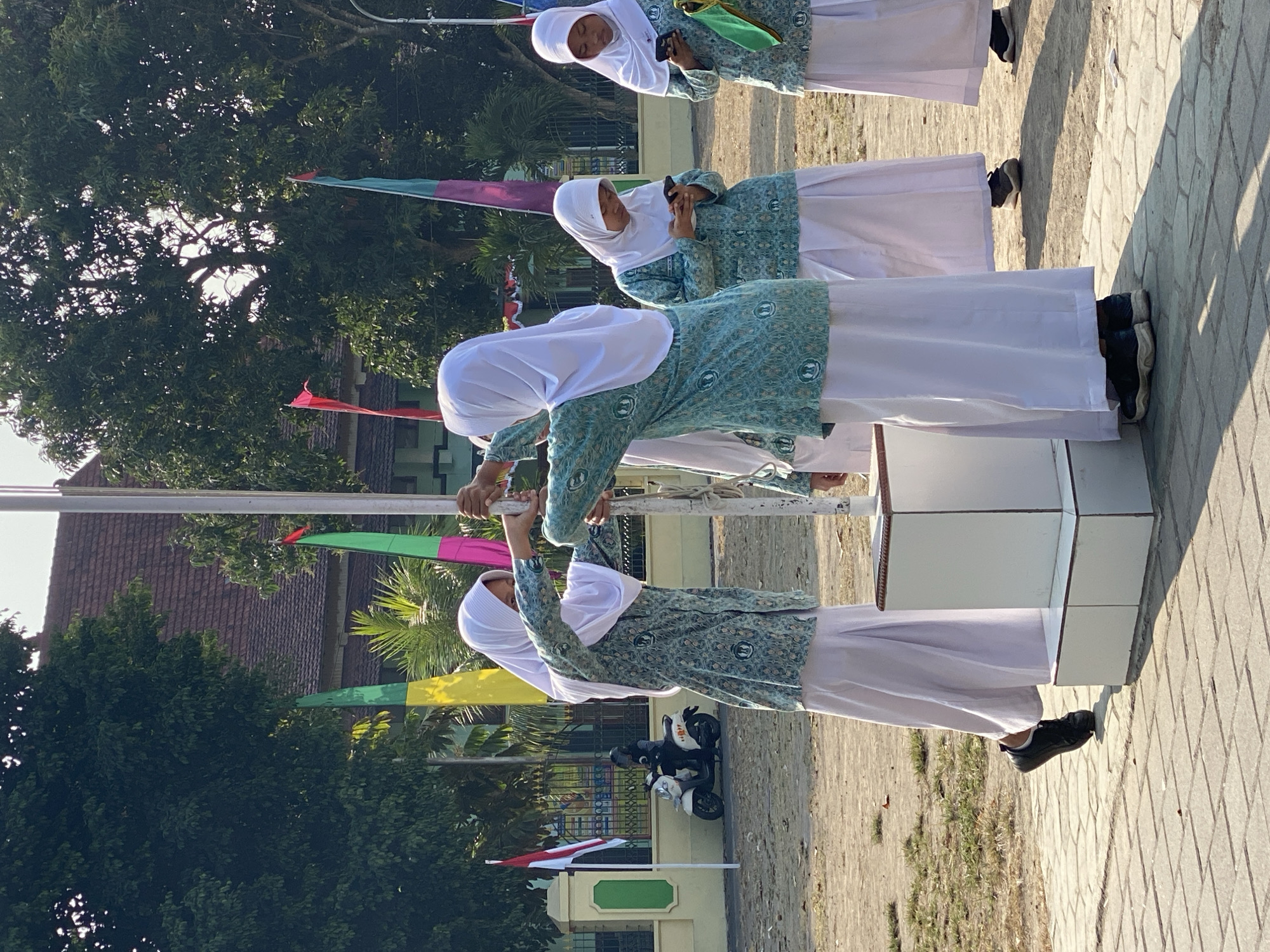 In Kasihan, Indonesia, on a bright, sunny day, four young women wearing matching teal patterned tunics and white hijabs are gathered around a flagpole base. Two women in the foreground are actively engaged, one on the left reaching up to adjust the flagpole rope, while the woman on the right assists her. Both are wearing long white skirts and dark sneakers. Behind them, two other women, also in similar attire, are looking down at their mobile phones. A white scooter with a rider wearing a helmet is parked to the left of the flagpole. In the background, a building with a tiled roof is visible, along with several colorful flags. The ground is a mixture of paved stones and dirt, with patches of dry grass.
