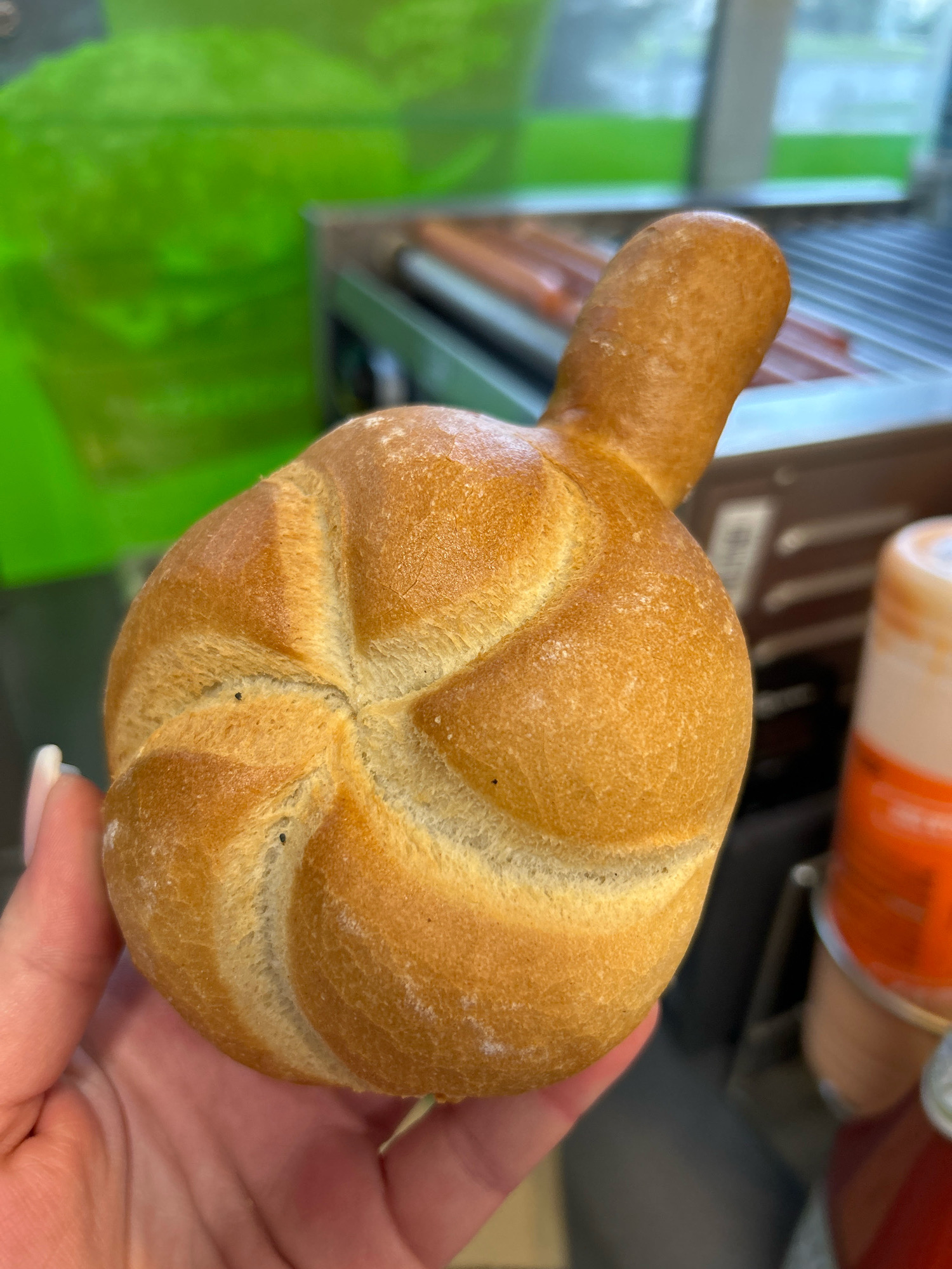 A close-up shot shows a person's hand holding a kaiser roll with a short, stubby appendage. The kaiser roll is golden brown and lightly dusted with flour. The background is blurred, but appears to be an indoor setting, possibly a food establishment. In the background, hot dogs are visible on a roller grill, and a red and white bottle is also present. The image does not provide any information about the location in Szczecin, Poland.