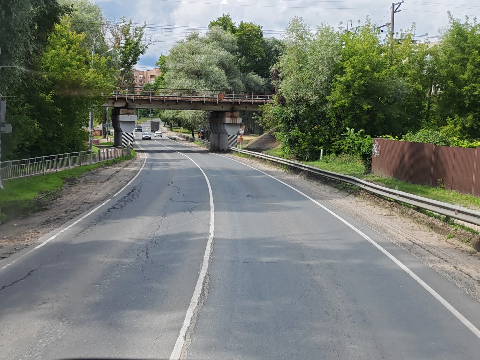 The image shows a road in Likino-Dulyovo, Russia, with a pedestrian bridge overhead. The road is asphalt with visible cracks and appears to be a two-lane road with white lane markings. There are trees and foliage on both sides of the road, and a metal guardrail runs along the right side. In the distance, a truck and several cars are visible on the road. The pedestrian bridge is made of metal and concrete, and appears to be old. The sky is cloudy. The environment suggests a suburban or semi-rural area within the city.