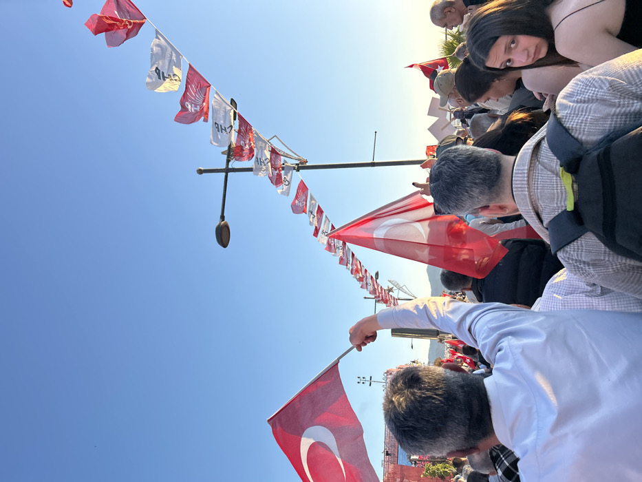 The image shows a crowd of people gathered in İzmir, Türkiye (38.437, 27.141), with a line of Turkish flags strung across a streetlamp. The people are looking towards something out of frame, and some are holding Turkish flags. One man is wearing a white shirt and raising his arm, holding a Turkish flag. He is in the foreground, while a man with a backpack is directly behind him. There is a young woman with long dark hair standing at the edge of the crowd, looking towards the camera. The sky is blue and the sun is shining.