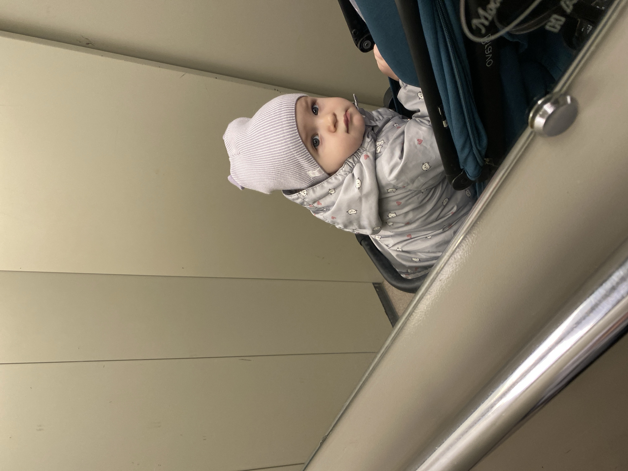 A baby wearing a striped gray and white beanie and a gray jacket with cat and heart patterns sits in a stroller. The stroller is teal and black, and a portion of its metal frame is visible in the foreground. The background consists of a beige wall with horizontal grooves. The setting appears to be indoors, possibly a public transportation vehicle, but the exact location in the city of Unknown, Unknown cannot be determined from the image.