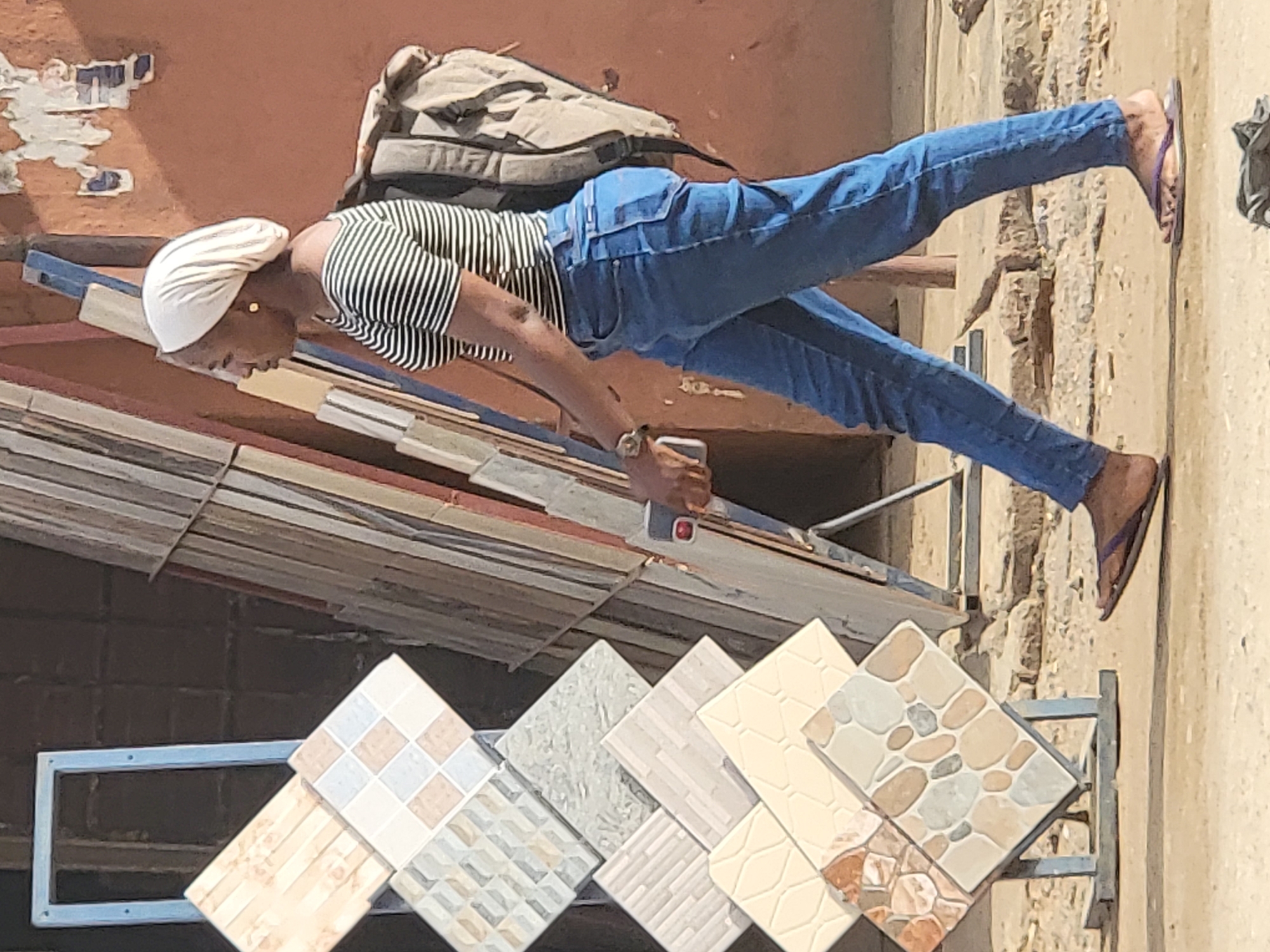 A medium, outdoor shot shows a woman in Maiburiji, Nigeria, levitating at an angle above a rack of decorative floor tiles. The woman has dark skin and wears blue jeans, a black-and-white striped top, a white head covering, and sandals. She holds a gray smartphone in her hand. A tan backpack rests on her back. The background is an orange brick wall, with faded, peeling paint and a beige stone wall. The bottom of the frame contains a metal rack with several decorative floor tiles in various earth-tone patterns. The image is taken in daylight with shadows.