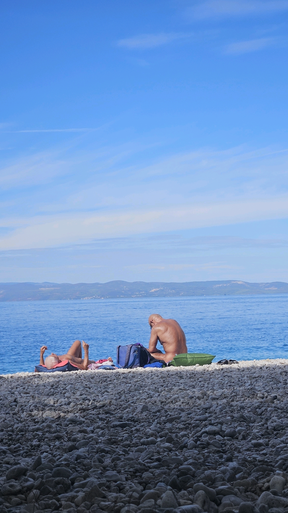 The image depicts a beach scene in Općina Podgora, Croatia, with two individuals sunbathing by the sea. In the foreground, the beach is covered in smooth, grey pebbles. In the mid-ground, a person lies on their back on a blue and orange mat, facing upwards with their legs bent and arms raised. Next to them, a blue duffel bag and some red fabric are visible. Further to the right, a bald individual sits on a green cushion, facing away from the viewer and towards the sea. This person is shirtless, with their back to the camera, and appears to be looking down at their hands. A blue backpack is situated behind them. The sea is a vibrant blue, extending to the horizon where it meets a range of distant, hazy hills under a clear blue sky with wispy white clouds. The overall environment suggests a peaceful, sunny day at the beach.