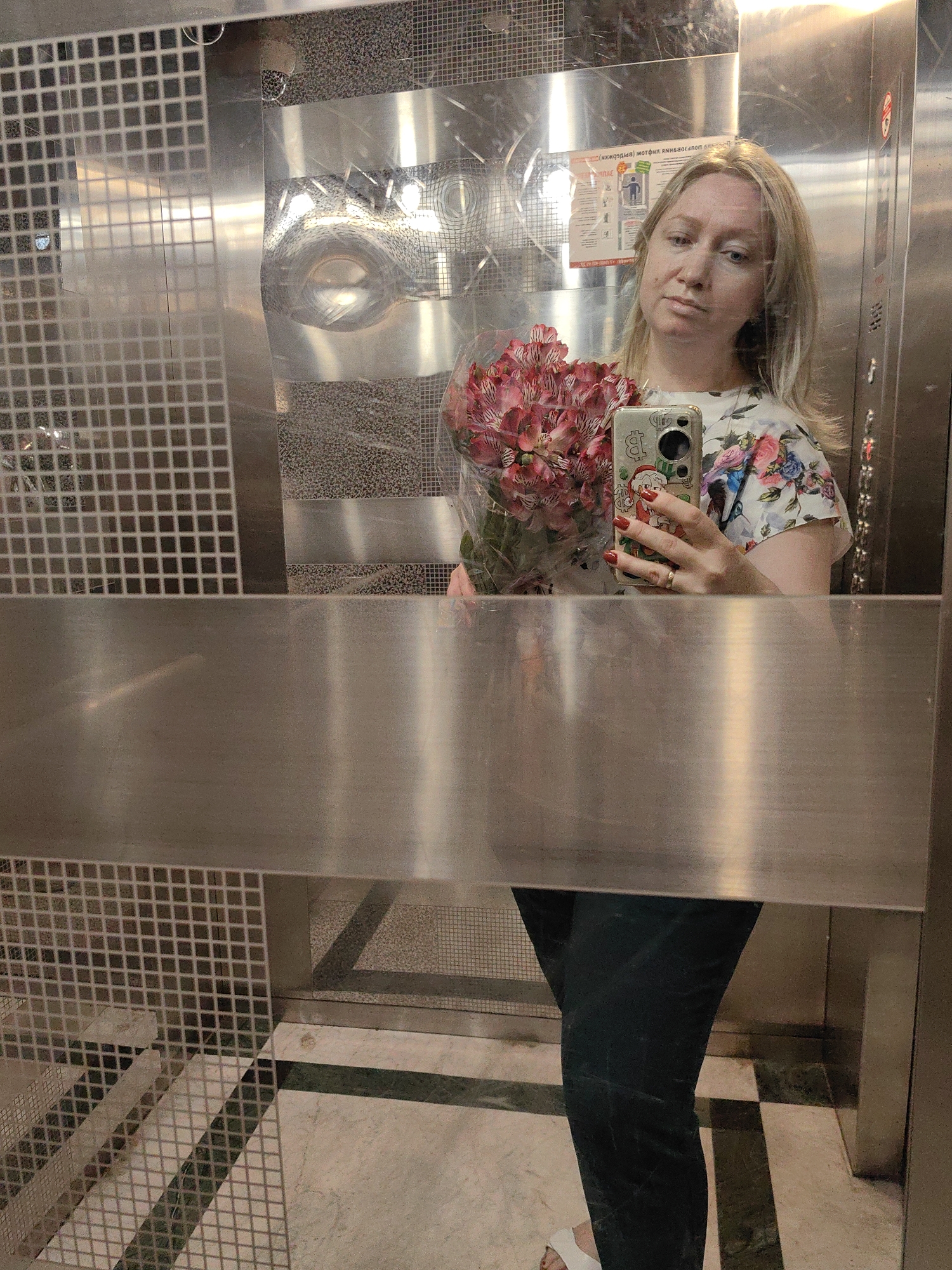 A blonde woman stands in an elevator, taking a selfie in the mirrored wall. She is holding a bouquet of pink and red flowers in her left arm and her smartphone in her right hand. The woman is wearing a white floral print blouse and dark pants. The elevator walls are made of brushed metal with a tiled pattern on the left side. The floor is marble with green accents. The image is captured in an elevator in Sochi, Russia.