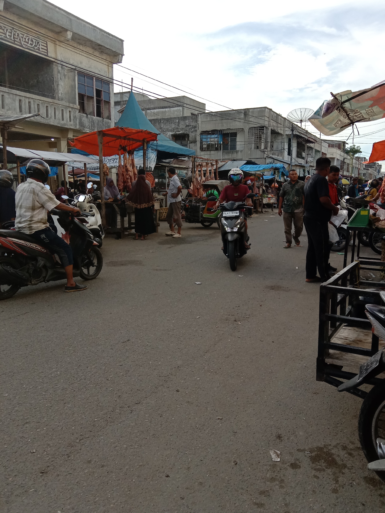 The image depicts a busy market scene in Lhokseumawe, Indonesia. A wide paved street serves as the main thoroughfare, with numerous vendors setting up stalls along the sides. People are seen walking, standing, and interacting with the vendors. Several motorcycles are present, some parked and others in motion, navigating through the crowd. Overhead, awnings and makeshift roofs provide shade. Buildings are visible in the background. Hanging meat is displayed at one vendor's stall. Overall, the scene conveys a sense of bustling activity typical of a Southeast Asian market.