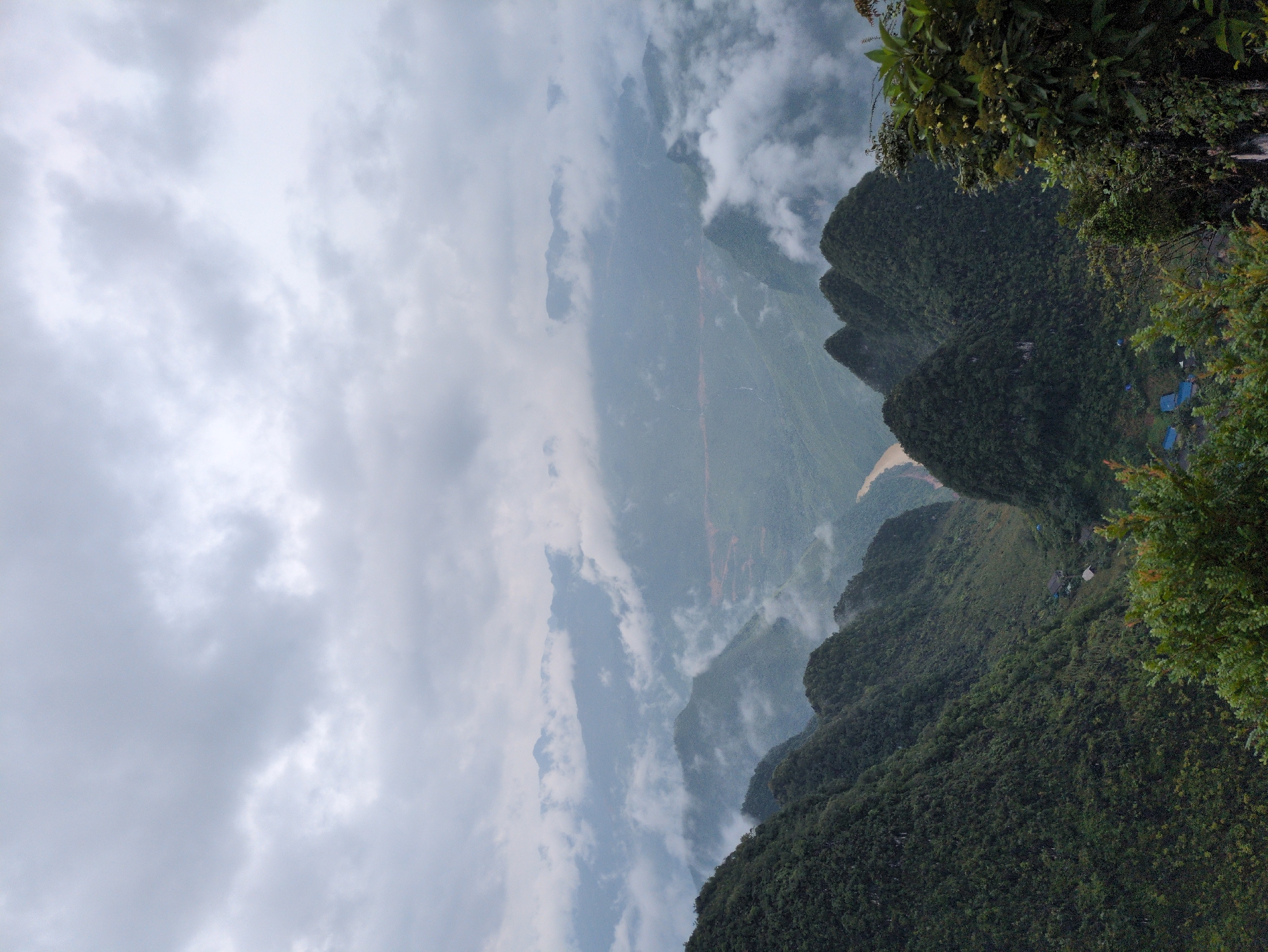 The image depicts a dramatic, misty mountain landscape in Đồng Văn Commune, Vietnam, characterized by lush, verdant peaks shrouded in low-hanging clouds. In the foreground, dark green, densely forested mountains dominate, with their slopes appearing steep and rugged. A winding, muddy-colored river snakes through a valley in the mid-ground, partially obscured by mist and the contours of the terrain. Small, blue-roofed structures, indicative of a village or settlement, are visible nestled within the valley floor. In the far distance, layers of receding mountain ranges fade into the atmospheric haze, creating a sense of immense depth. The sky is overcast with thick, gray clouds, contributing to the diffused light and ethereal quality of the scene. The overall impression is one of a remote and wild natural environment, likely captured during or after rainfall, given the prevalent mist.
